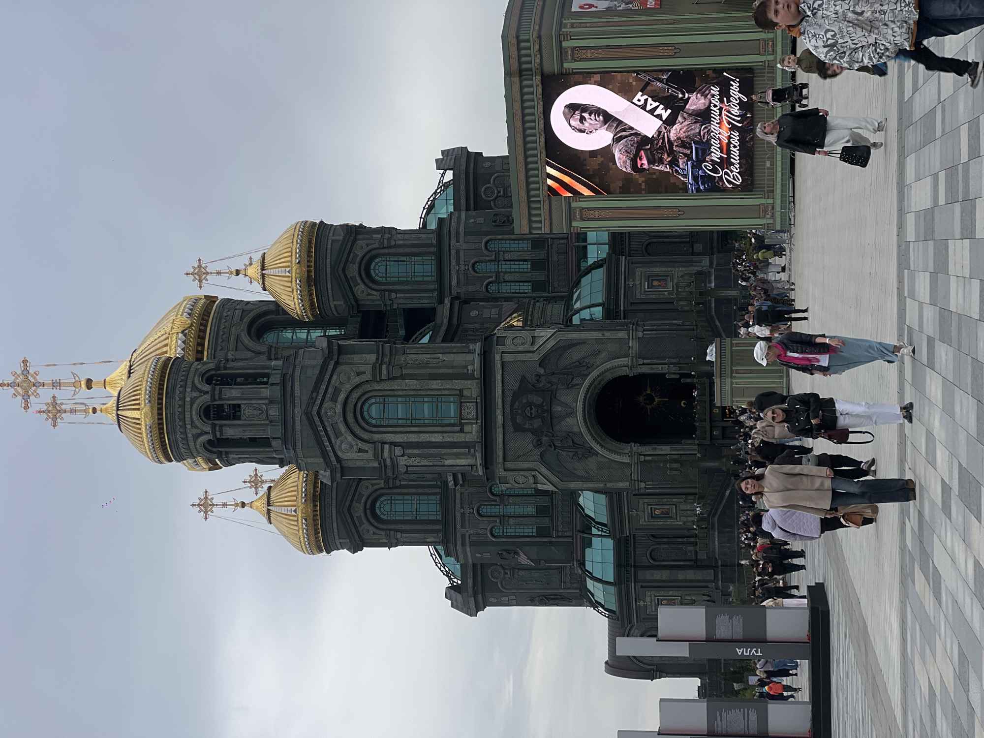 The image depicts the Main Cathedral of the Russian Armed Forces in Zelenaya Roshcha, Russia. The cathedral has multiple golden domes and a dark-colored, ornate exterior. A large video screen is visible on an adjacent building, displaying an image of a soldier and the text "9 MAY," likely referring to Victory Day. A crowd of people is gathered in the plaza in front of the cathedral. The ground is paved with light and dark gray bricks. The sky is overcast. There are informational displays in the foreground. People are seen walking and standing around the plaza.
