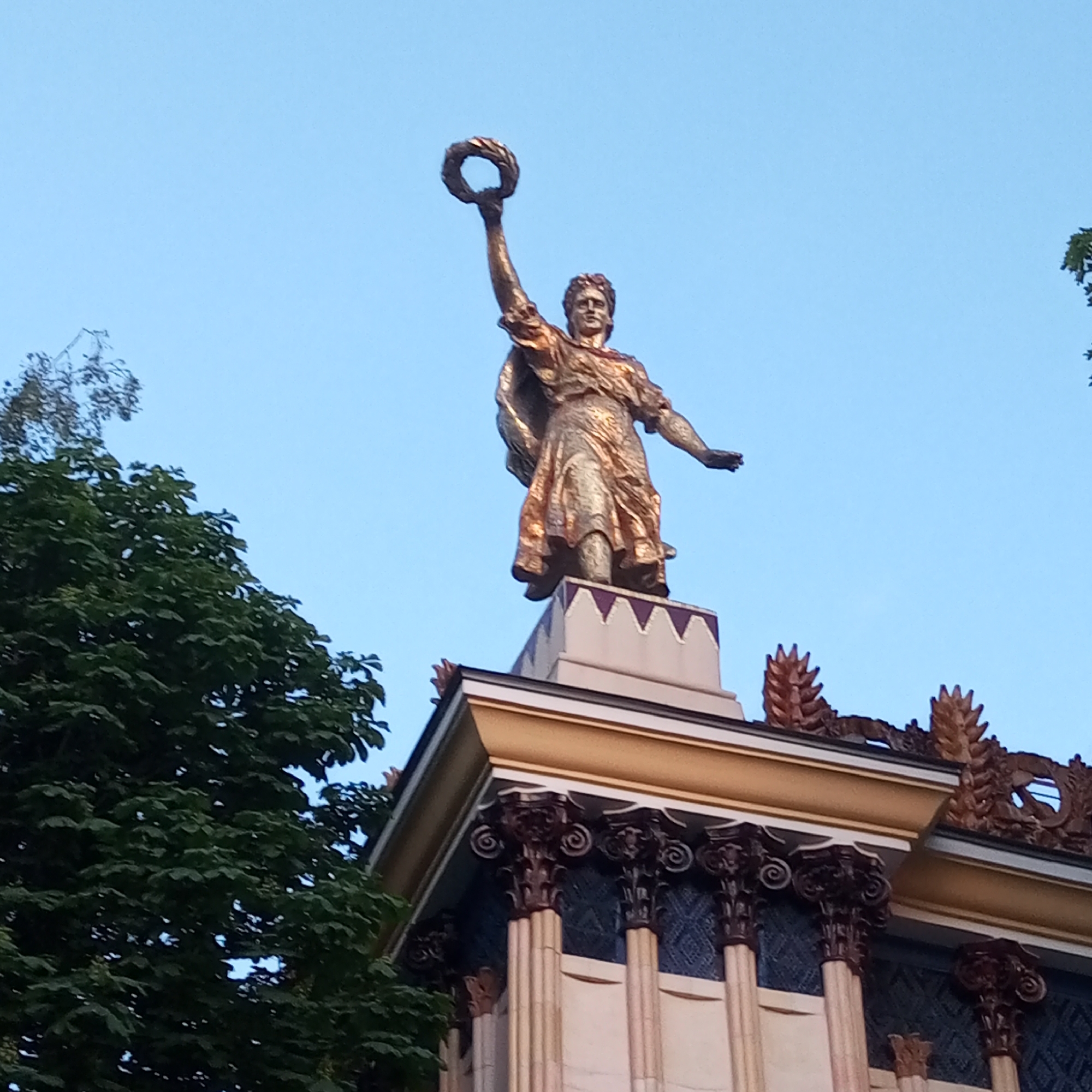 A gilded statue of a female figure stands atop a decorative architectural structure under a clear blue sky. The statue, depicting a woman in classical-style flowing robes, raises a laurel wreath high overhead with her right arm, while her left arm is slightly extended forward. She rests on a light-colored pedestal accented with purple triangular motifs. The building below features a prominent golden-yellow cornice and a series of dark, ornate columns with intricate capitals, set against a background of dark blue patterned wall panels. Stylized leaf or flame-like decorative elements adorn the roofline. Lush green tree foliage partially obscures the lower left portion of the building and the sky. This scene is located at the VDNKh (Exhibition of Achievements of National Economy) complex, specifically on the Central Pavilion (Pavilion No. 1), in Moscow, Russia.