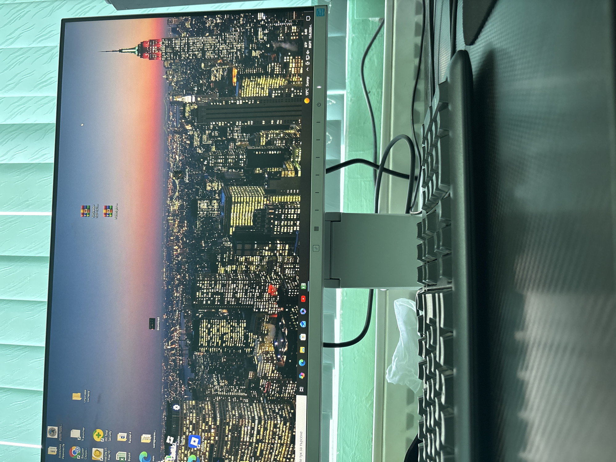 The image shows a computer monitor displaying a nighttime cityscape with the Empire State Building illuminated in red and green. The desktop features various icons for applications such as "Roblox Player," "TLauncher," "Razdel 4," and "FullBright." The operating system appears to be Windows, indicated by the taskbar at the bottom of the screen which includes icons for Microsoft Edge, File Explorer, and other applications. The bottom right corner of the taskbar displays the date "15.6.2026 г." and the time "8:49". The environment is a desktop setup with a keyboard in the foreground, a tissue box behind the keyboard, and a green-tinted blind or curtain in the background. There are no people visible in the image. The location in Svoge, Bulgaria is not evident from the image content; the cityscape is clearly New York City.