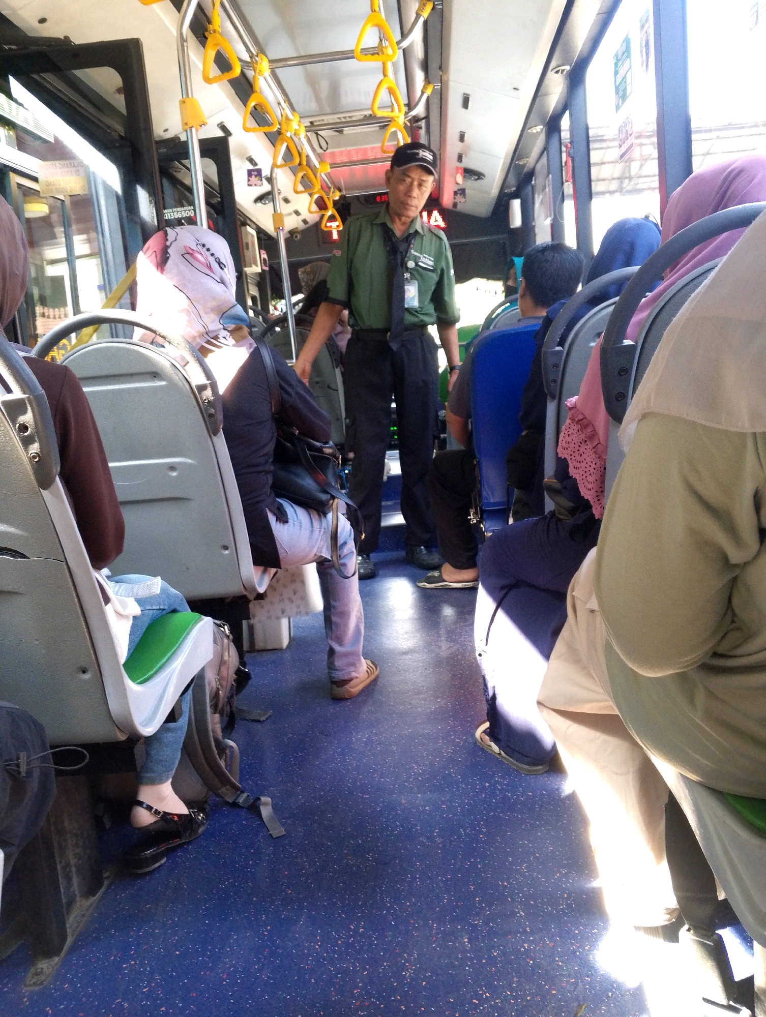 The image depicts the interior of a bus in Meri, Indonesia. The bus is occupied by several passengers and a driver. The driver, an older man wearing a green uniform and a black cap, stands near the front of the bus. Passengers are seated on the right side of the bus, with their backs to the camera. One passenger in the foreground is wearing a white hijab with a floral pattern. The bus floor is blue with red and white speckles. Yellow handrails are visible along the ceiling of the bus. Sunlight streams in from the right side of the bus, creating bright highlights and shadows.