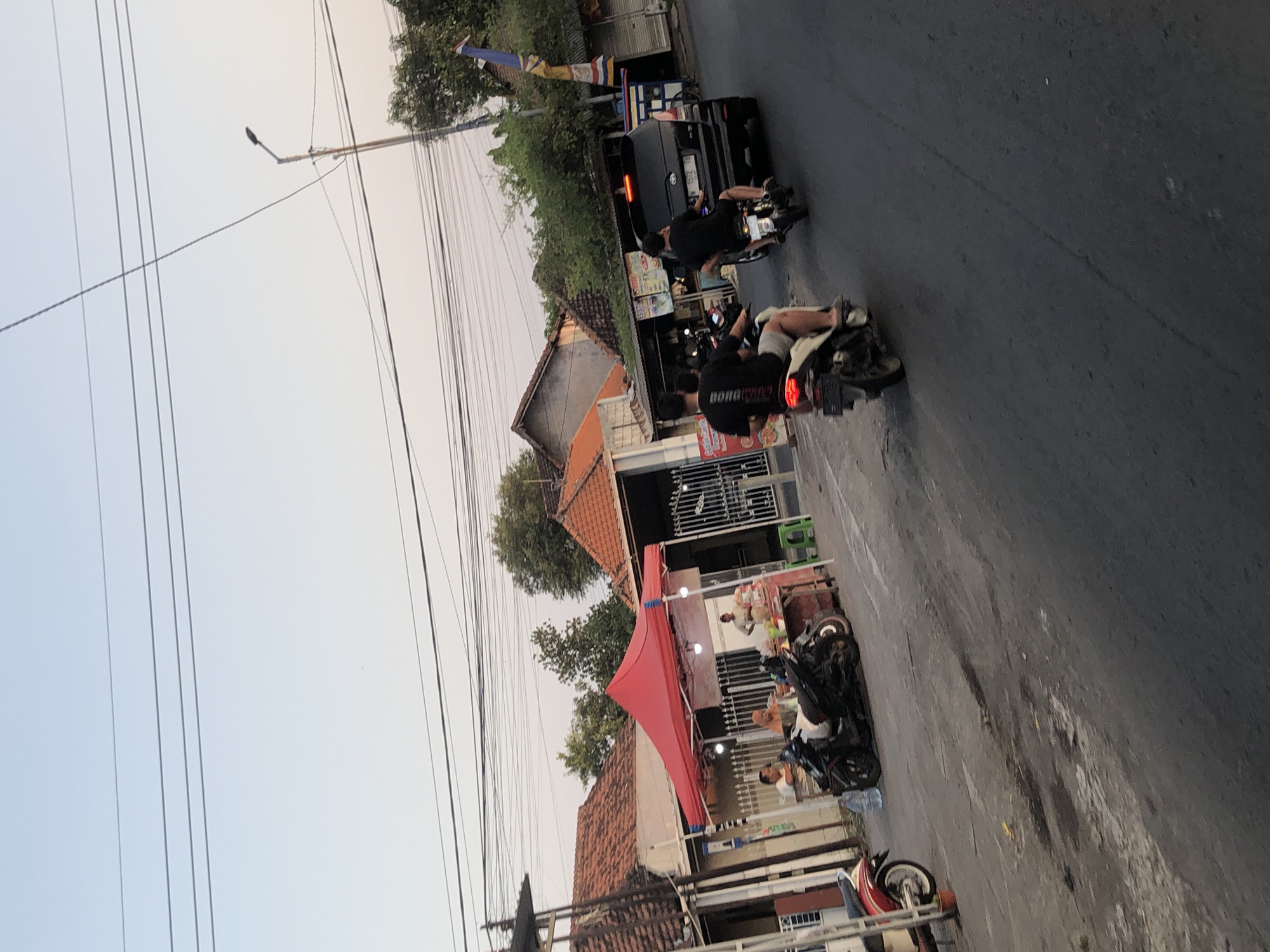 This is a full, eye-level, outdoor shot captured in Glagah Arum, Indonesia, at dusk. The scene depicts a street with several buildings lining one side, featuring traditional red-tiled roofs. In the foreground, the asphalt road is visible, with various motorcycles parked and in motion.
On the left side of the street, a red tent is set up, beneath which several people are seated at a makeshift food stall. A motorcycle is parked in front of the tent. Further down the street, a dark-colored van is visible, and two men on motorcycles are riding past it. One of the motorcycles is in the immediate foreground, traveling away from the viewer.
The environment appears to be a local commercial area or a roadside market. Overhead, a network of power lines crisscrosses the sky, with a street lamp visible on the right. The sky is clear and exhibits the soft hues of twilight. The overall impression is that of a casual, active street scene in a residential or commercial district.