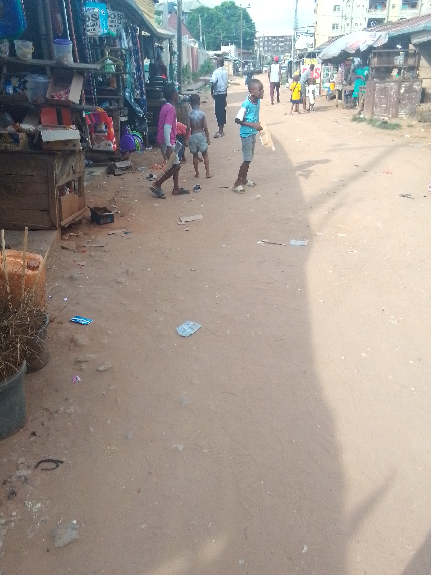 The image depicts a street scene in an urban area, likely a market or commercial district. The ground is dusty and littered with trash. Several children are present in the foreground, some partially clothed. Adults are visible further down the street, some walking and some standing near makeshift stalls or shops. The buildings in the background appear to be residential and commercial structures. The scene conveys a sense of everyday life in a developing area.