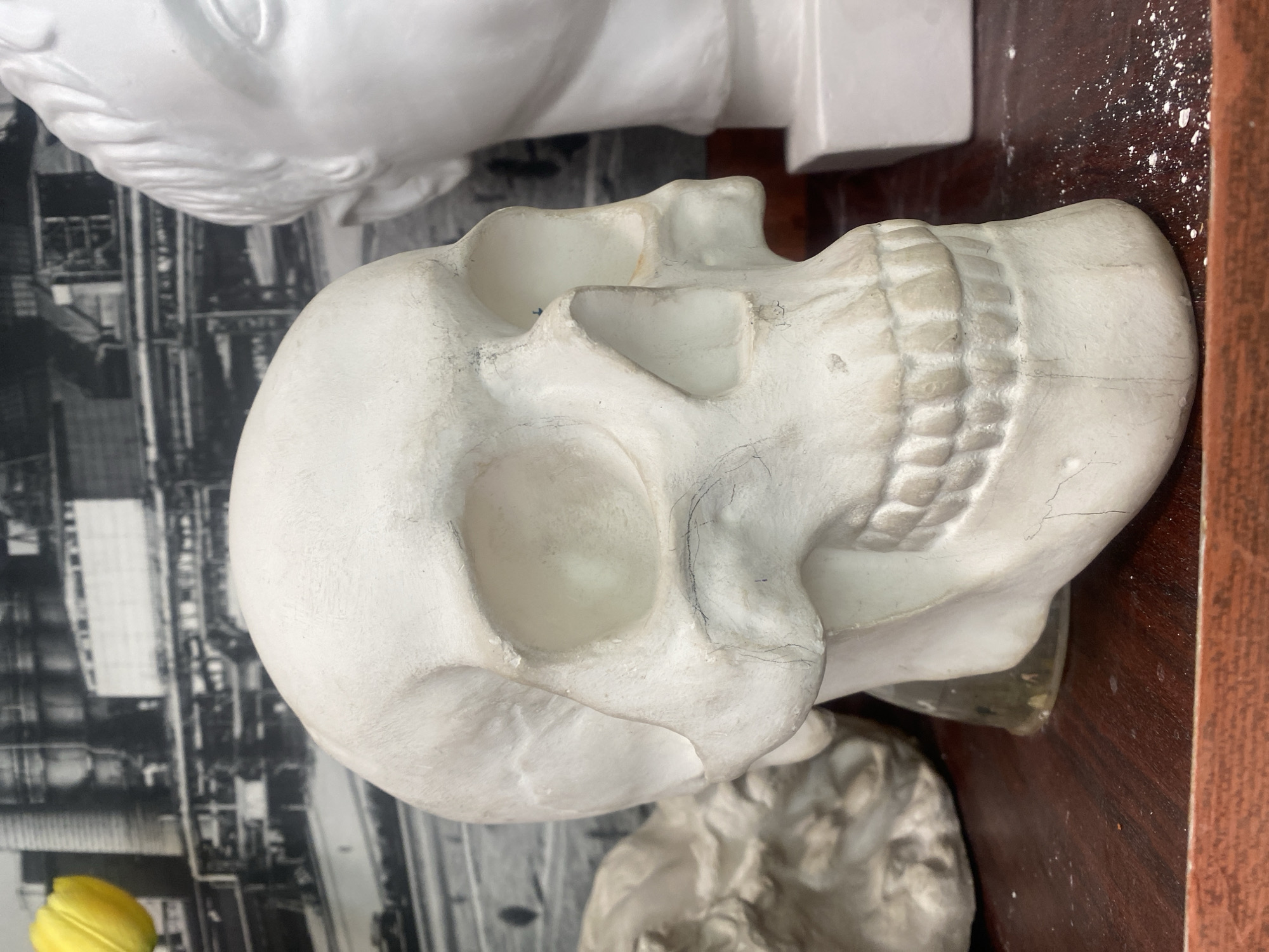 The image shows a white plaster skull resting on a brown wooden surface. The skull is positioned in the center of the frame, angled slightly to the right. The background includes a blurred black and white print depicting industrial buildings. Another white plaster bust is partially visible above and behind the skull. A small yellow object is visible on the left edge of the image. The surface is splattered with white powder. The location is impossible to determine from the image.