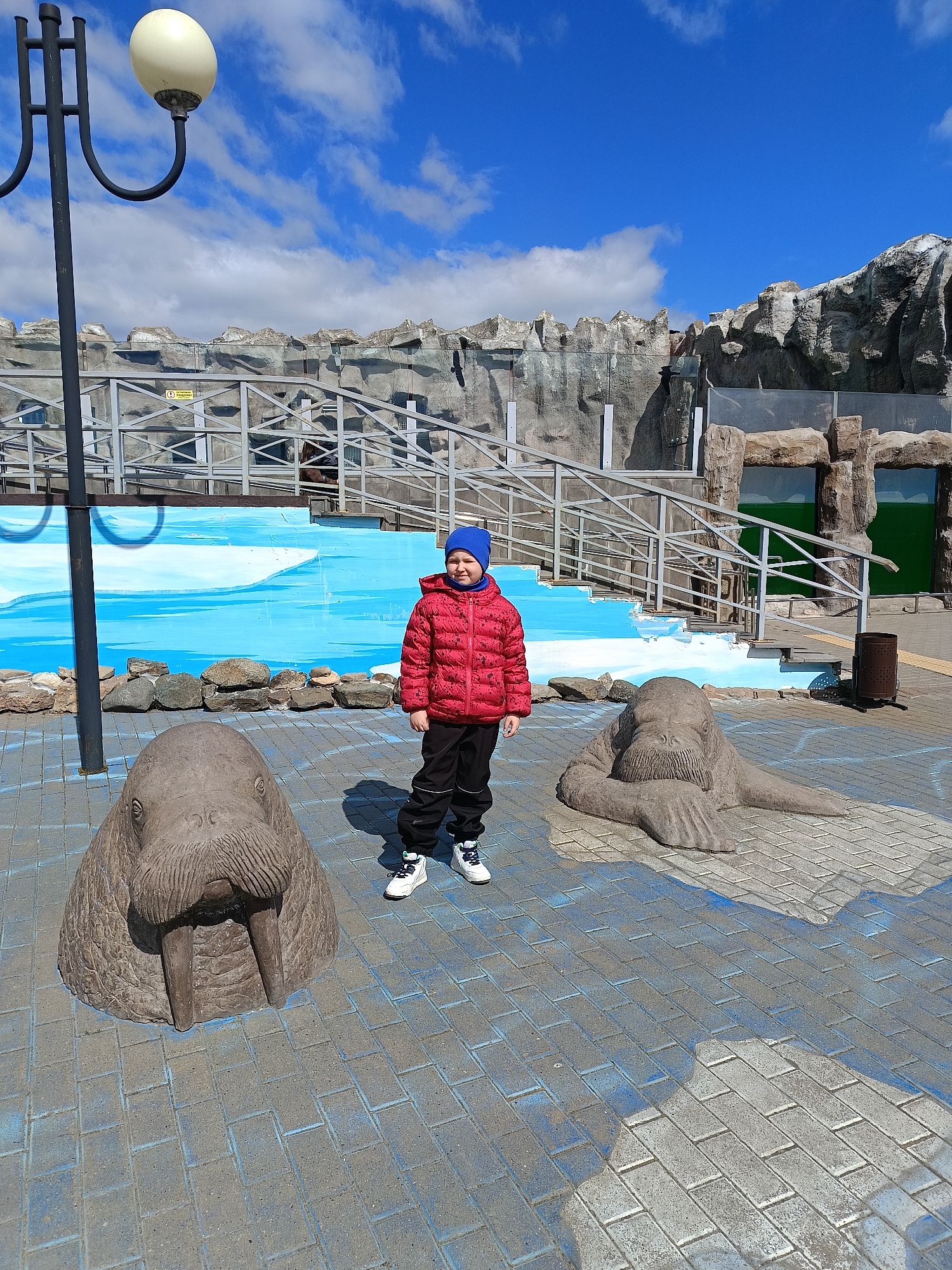 A child stands between two walrus statues in Izhevsk, Russia. The child, wearing a red jacket and blue hat, is centered in the frame. The statues are on a blue-painted brick surface. Behind them is a painted mural of an arctic landscape and a walkway with metal railings. The sky is blue with scattered clouds. A lamppost is visible on the left.