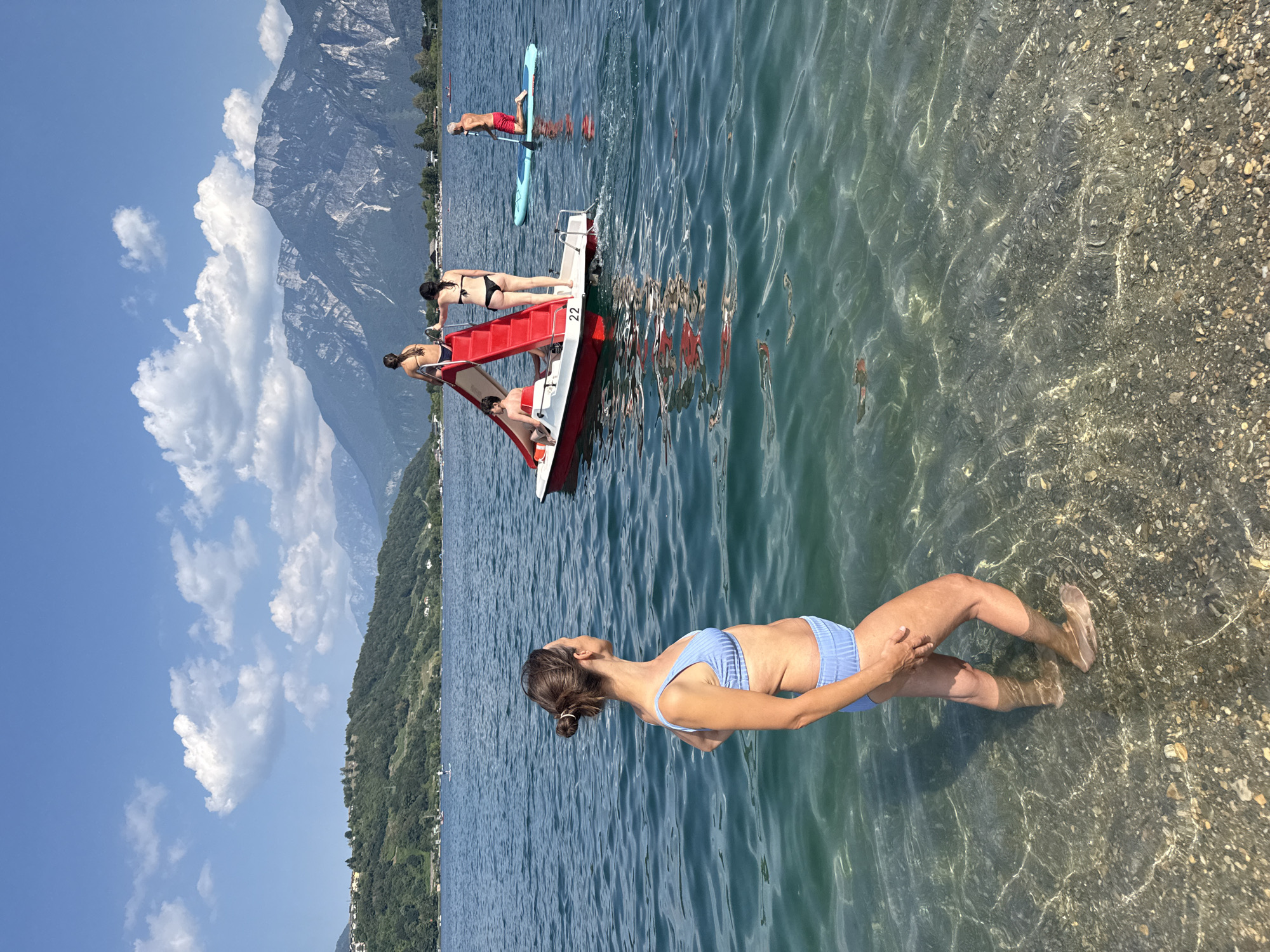A woman in a blue bikini stands in shallow, clear water at the edge of a lake, facing away from the camera and looking towards the water. In the middle ground, a red and white pedal boat with a slide is occupied by several people. One person is on the slide, another is standing at the top of the slide, and a third is sitting on the boat. Further in the background, a person on a paddleboard kneels in the water. The lake is surrounded by lush green hills and majestic mountains under a blue sky with scattered clouds. The scene is set in Calceranica al Lago, Italy.