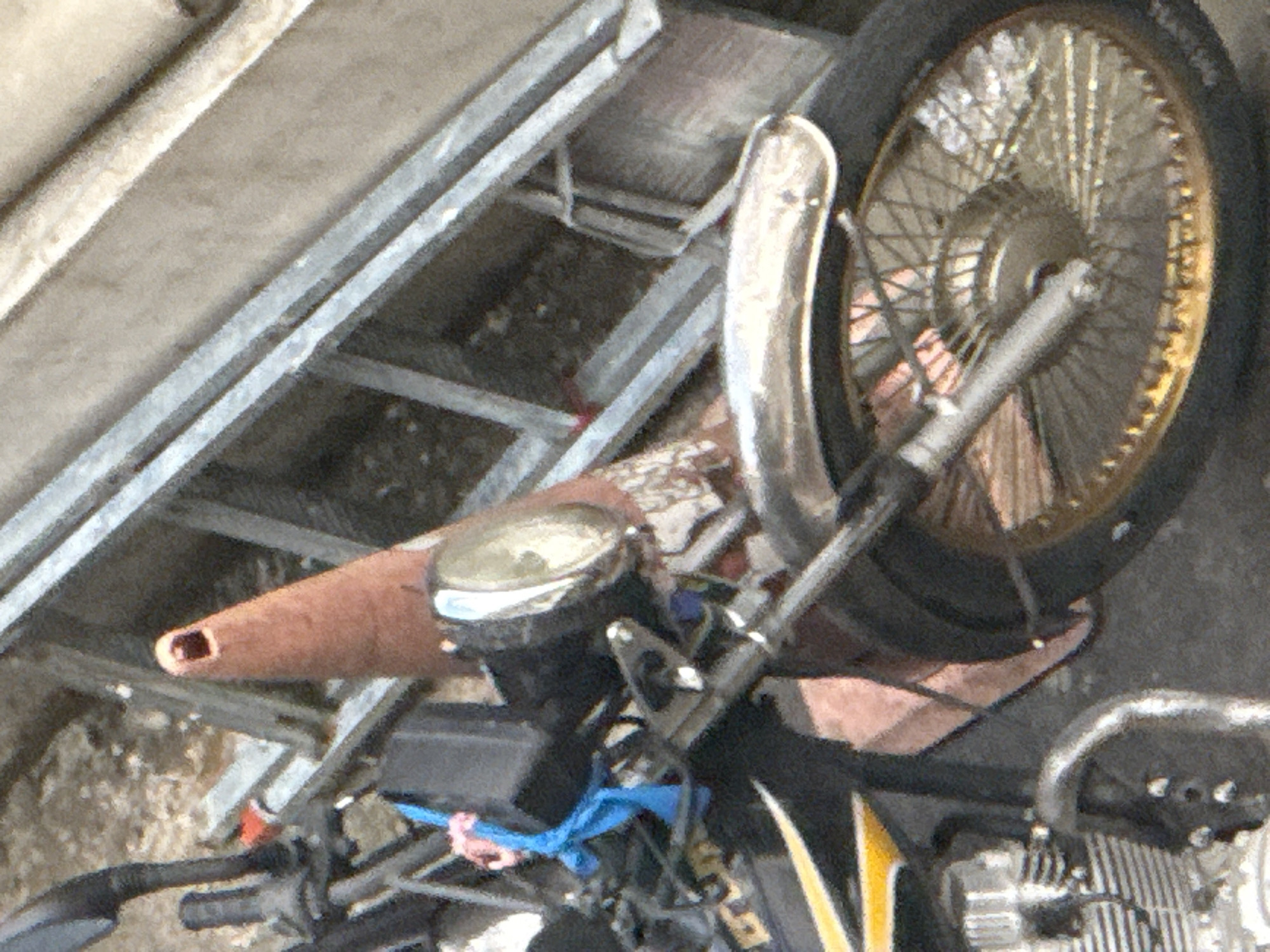 The image shows a close-up view of a motorcycle parked outdoors, possibly in Tbilisi, Georgia, as suggested by the presence of what appears to be a construction ladder in the background. The motorcycle has a metallic grey body with yellow accents. A brown traffic cone is positioned near the handlebars, which are black. A bright blue ribbon or tie is visible, attached to the motorcycle's controls. The front wheel and part of the engine are also visible. The ground appears to be a rough, outdoor surface.