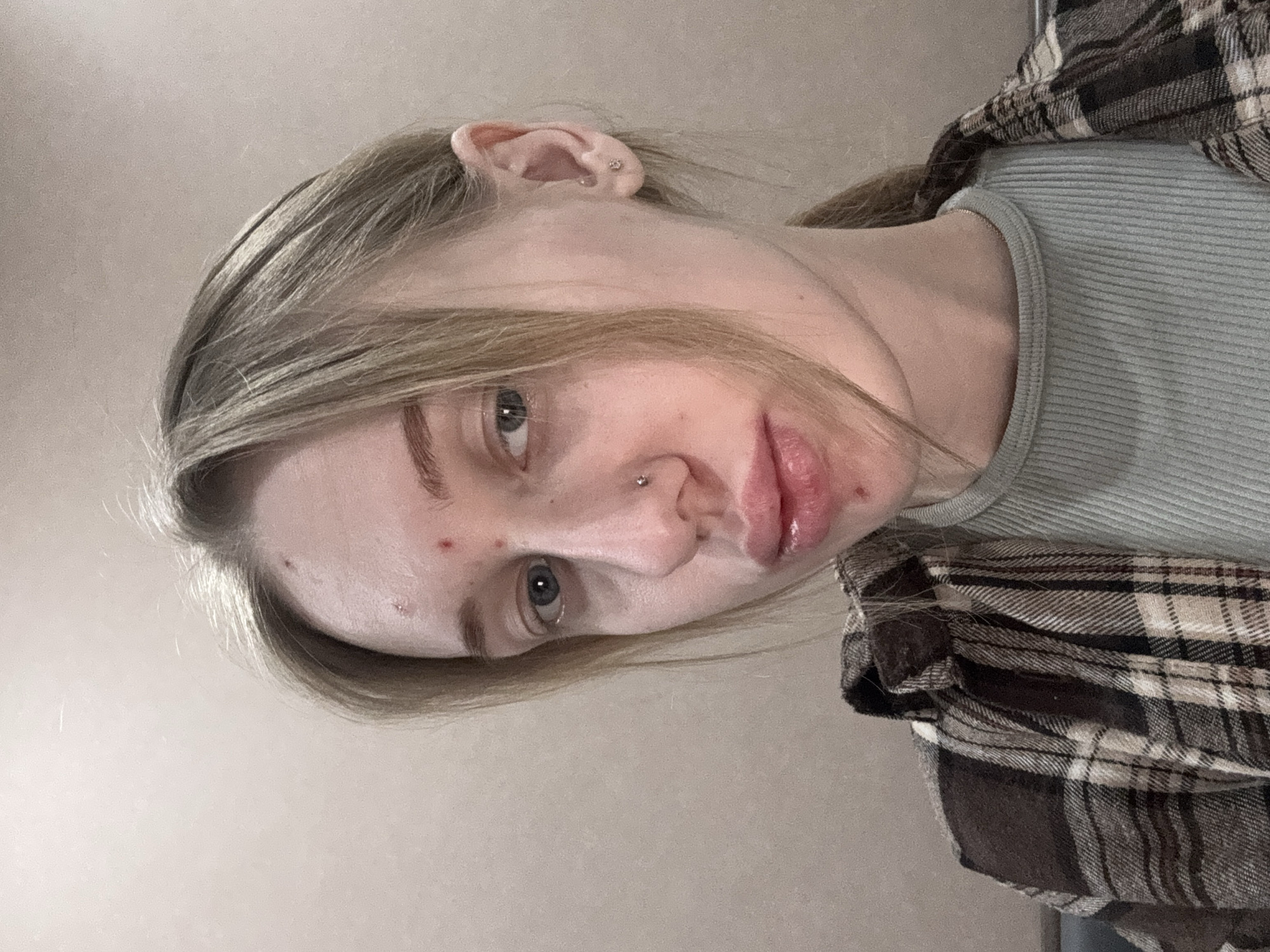 A photograph depicts a woman with light blonde hair and fair skin, facing the camera at a tilted angle. She has a nose piercing and what appears to be a small blemish on her forehead. She is wearing a light green top and a plaid shirt. The background is a plain, light-colored wall. There is no indication of the location being Novoalexandrovka, Russia, or any other specific location.