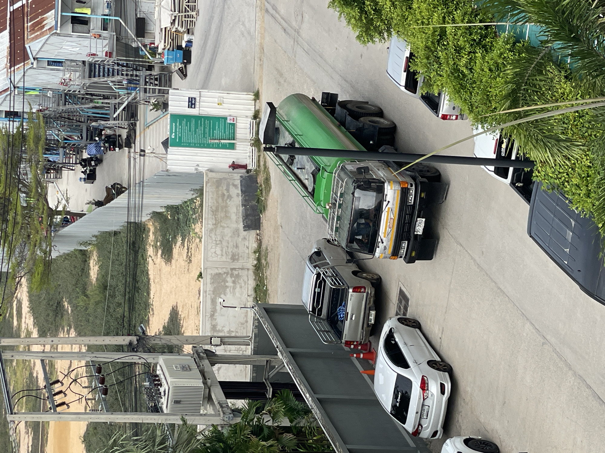 The image captures an outdoor scene in Pattaya City, Thailand, focusing on a street or industrial area. Dominating the foreground is a large green tanker truck with a silver cab, parked on a paved surface. To its left, a silver pickup truck is parked, and further left, a white sedan is visible. An orange traffic cone is positioned near the pickup truck. In the background, there are buildings with outdoor staircases, parked motorcycles, and what appears to be a construction or industrial site with fencing and stacked materials. Electrical infrastructure, including poles, transformers, and wiring, is prominent on the left side of the image. Lush green foliage is present throughout the scene, particularly in the foreground and to the right. The overall impression is one of a functional, possibly industrial, area in an urban setting.