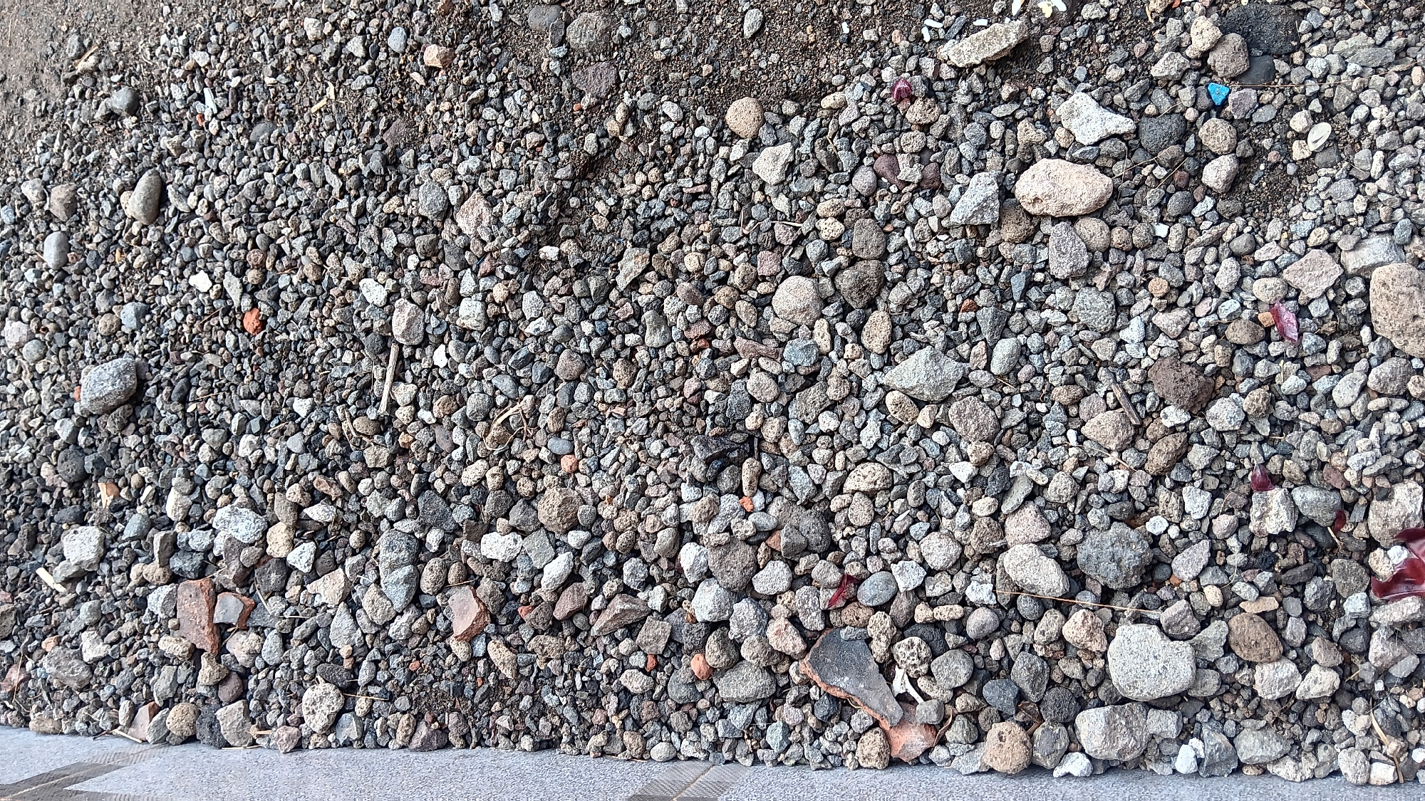 This image depicts a close-up, aerial view of a ground surface covered in a dense layer of small to medium-sized stones and gravel. The stones vary in color, ranging from light grey and white to dark grey and brown, with some appearing to be broken pieces of brick or pottery. There is also some finer dirt and sand mixed in with the larger stones, and a few dry twigs are visible. On the far left edge of the image, a textured, light grey concrete or stone border runs vertically. The scene is consistent with an outdoor area, such as a pathway, yard, or construction site, likely in an urban or suburban environment. The location is identified as Sonobekel, Indonesia. No people or specific actions are visible.