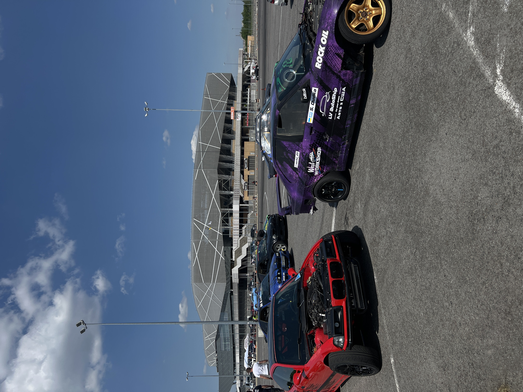 The image depicts a scene at the Arena Lviv stadium in Lviv, Ukraine. Several cars are parked in a lot in front of the stadium. A red car and a purple car are most visible with their hoods open, and other cars are parked behind them. The cars feature sponsor stickers like "ROCK OIL", "LV AutoShip", and "West Workshop". People are standing around some of the vehicles. The sky is clear with some scattered clouds, and light poles are visible.