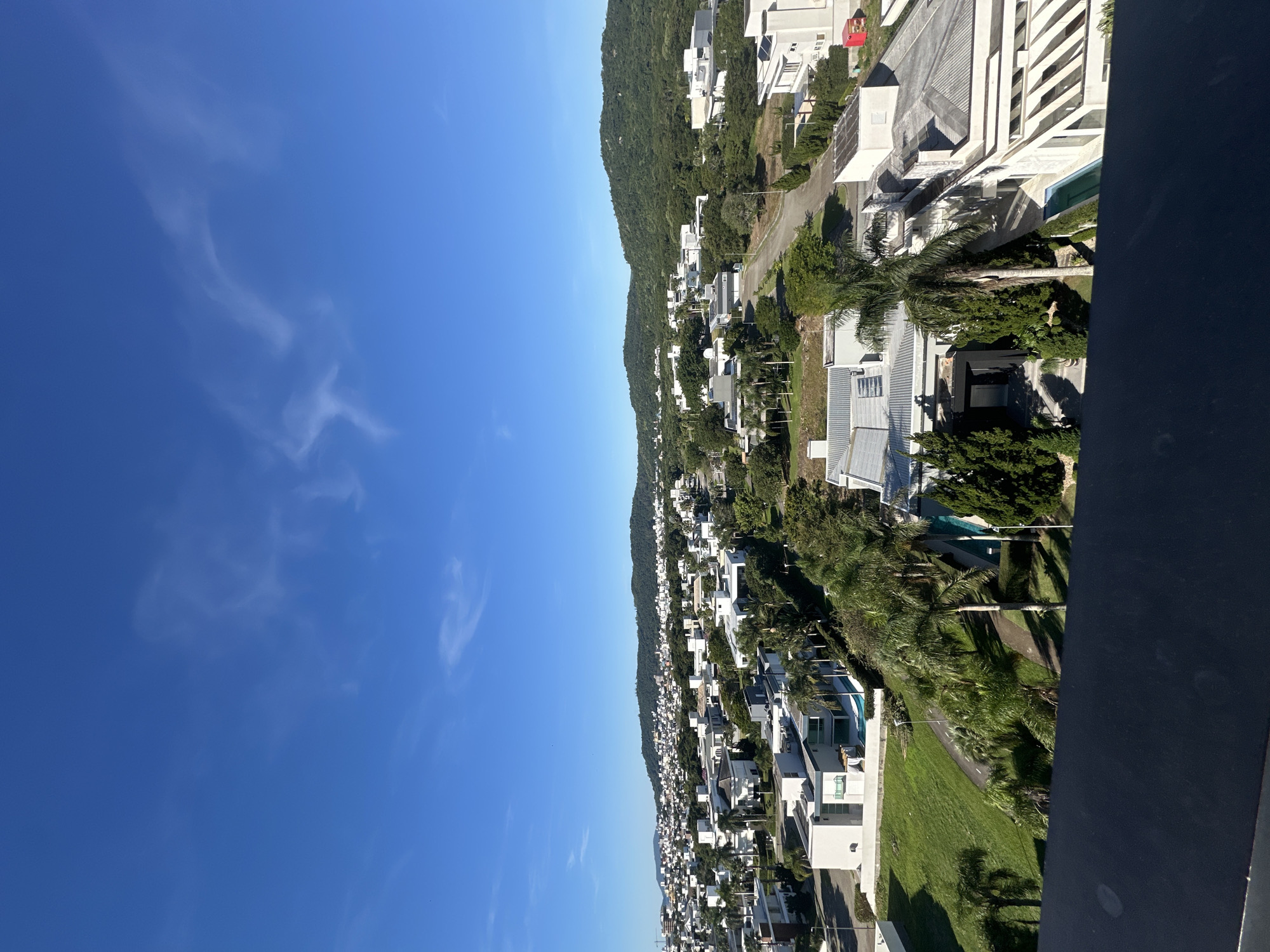 An aerial shot captures a bright, sunny day in Florianópolis, Brazil, with a clear blue sky dotted with wispy white clouds. The image showcases a dense residential area characterized by modern, white-washed houses with flat roofs, interspersed with lush greenery and palm trees. The houses are of varying sizes and architectural styles, some featuring swimming pools. In the background, rolling green hills covered in dense vegetation form a natural backdrop to the urban landscape. A road winds through the neighborhood, connecting the houses and hinting at a well-planned community. The foreground features a dark railing, suggesting the vantage point is from a balcony or rooftop. The overall impression is one of a prosperous and well-maintained residential neighborhood in a tropical or subtropical climate.