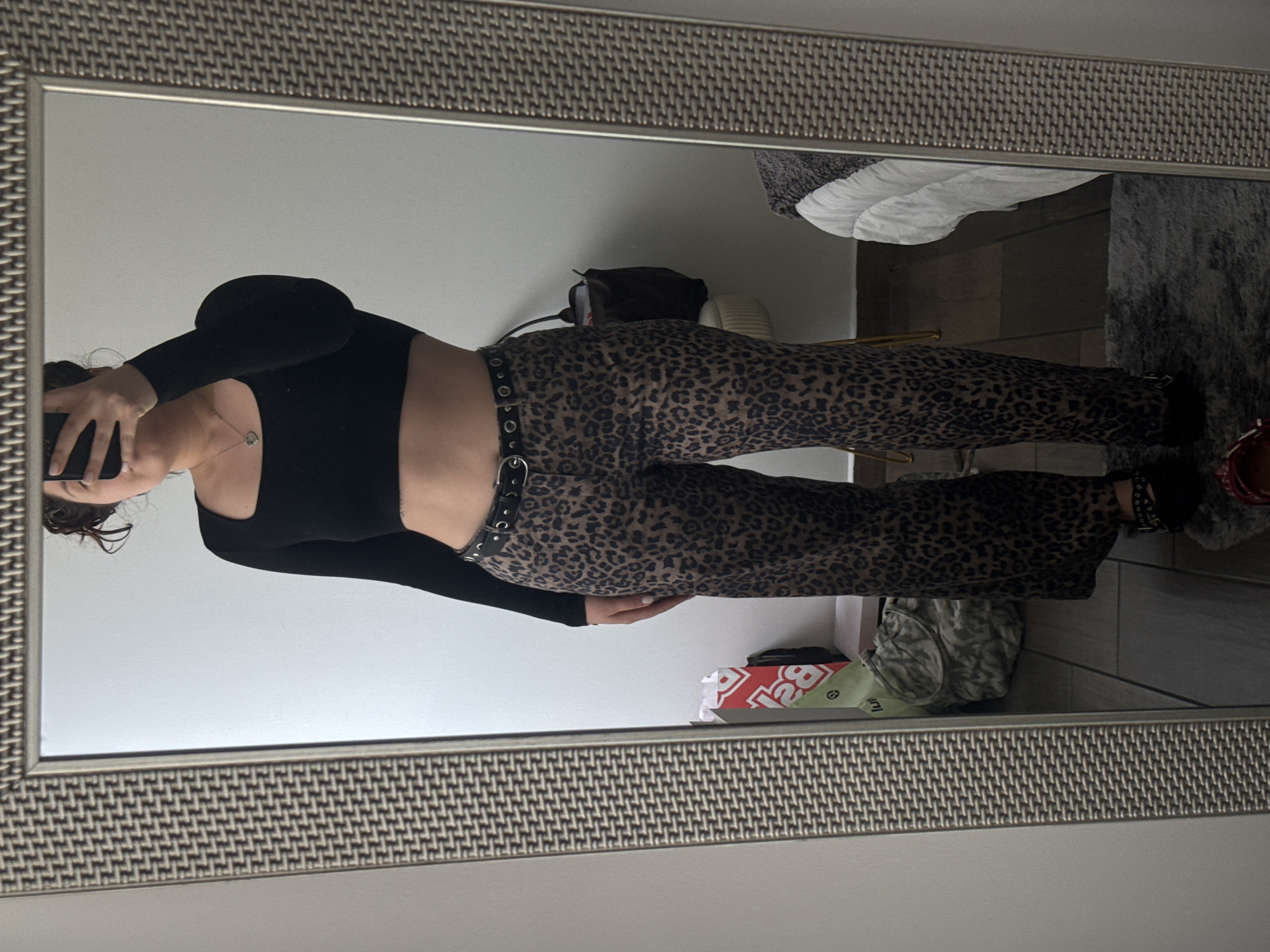 A young adult female is seen in a mirror selfie. She is wearing a long-sleeved black top and leopard-print pants with a belt. The environment reflected in the mirror includes a bed, a rug, and some items on the floor, including a shopping bag. The location and city cannot be determined.