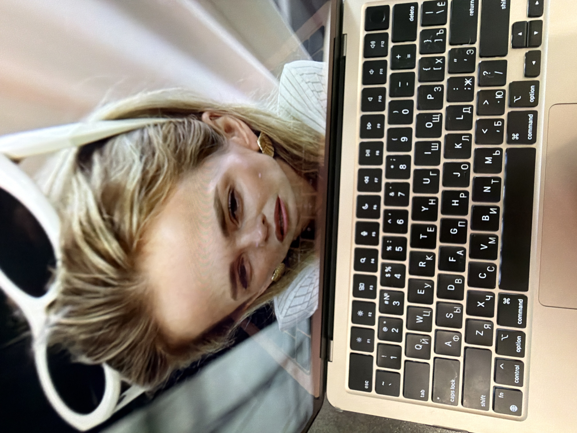 The image shows a laptop screen reflecting a woman with blond hair, sunglasses, and a white striped shirt. The laptop's keyboard is visible, with keys featuring both English and Cyrillic characters. The focus is on the laptop and its reflection, not on a specific location or city like Birobidzhan, Russia. There is no information in the image to indicate the location.