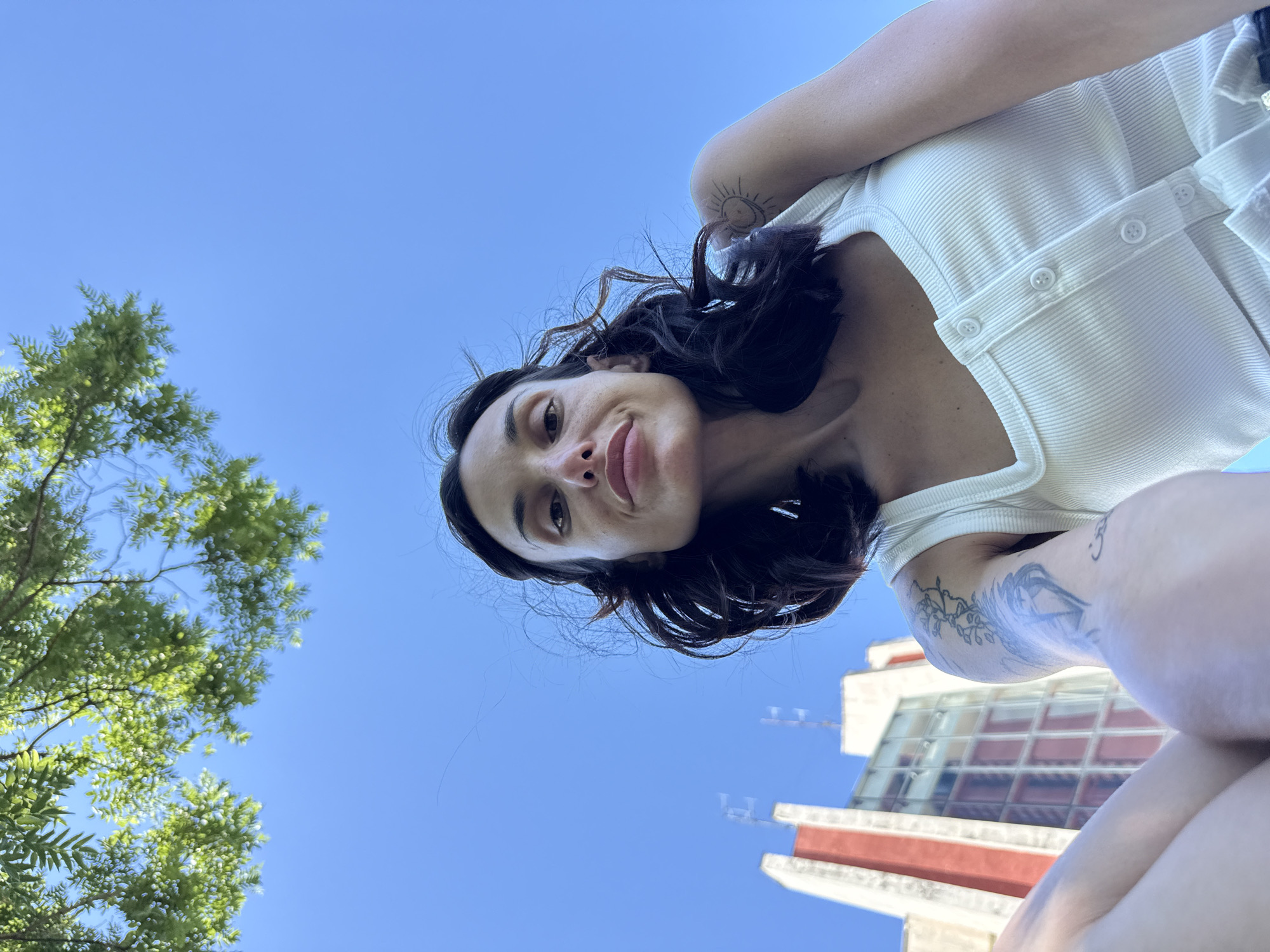 A low-angle, wide shot captures a young woman with dark, wavy hair, taken from a ground-level perspective looking upwards. She is wearing a white ribbed tank top with a square neckline and button detailing. Tattoos are visible on her left arm, including a sun and a floral design. Her gaze is directed upwards towards the camera, with her lips slightly parted.
In the background, a clear blue sky dominates the upper portion of the image. Below the sky, a building with distinctive red and white architectural features is visible, possibly an apartment block or office building, which is consistent with the urban environment of Pleven, Bulgaria. Green foliage from a tree hangs into the frame from the top left. The overall impression is a casual, outdoor portrait set against an urban backdrop.