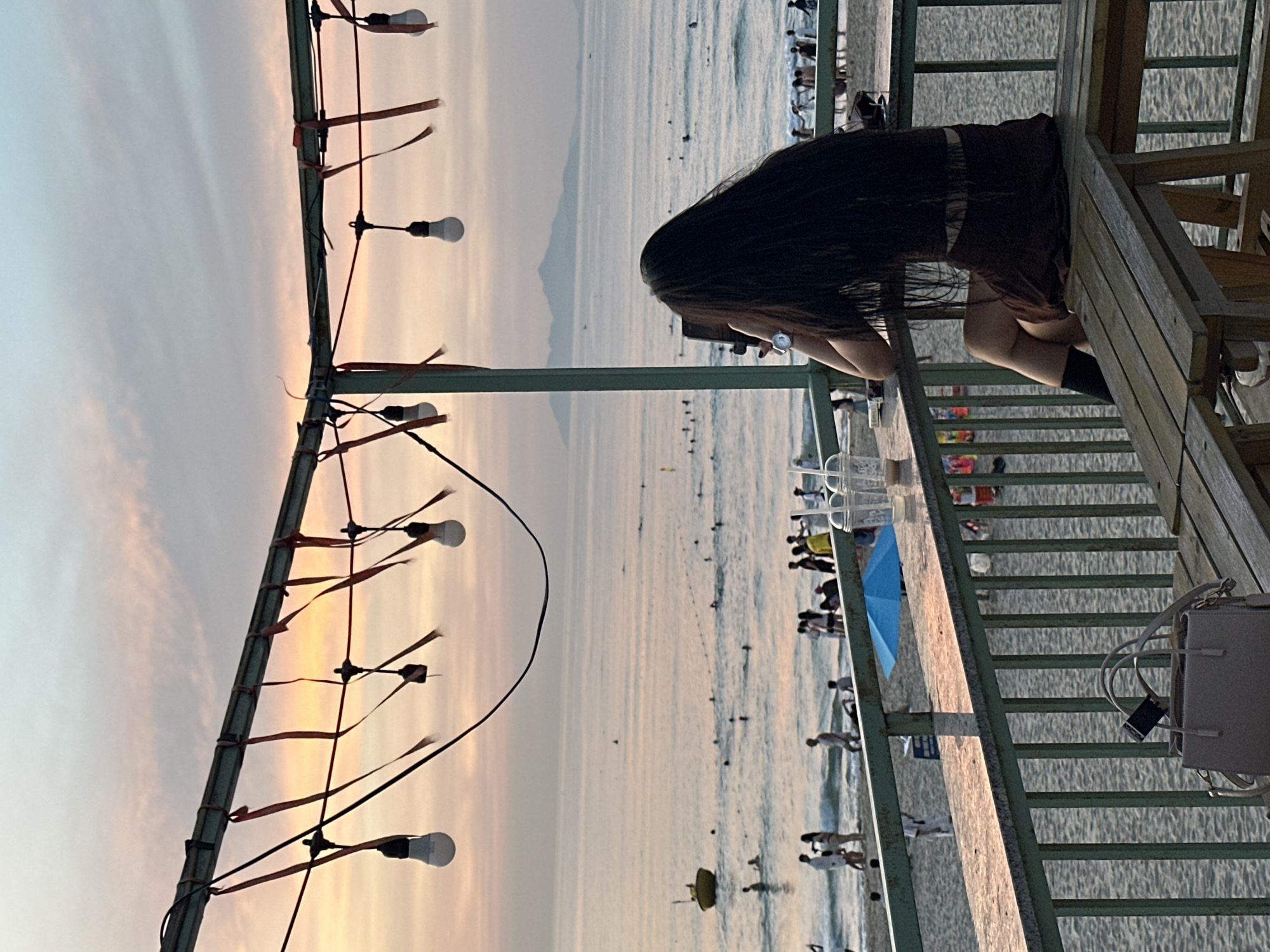 A young woman with long, dark hair sits at a table on a balcony overlooking a beach at dusk in Đà Nẵng, Vietnam. She is wearing a brown skirt and black socks. In front of her is a tablet computer, which she appears to be holding. A light-colored handbag rests on a wooden bench beside her. The balcony is strung with lights and decorated with red ribbons. The beach is dotted with people, some swimming in the ocean and others walking along the shore. The sky is a soft gradient of pink, orange, and blue, with distant mountains visible on the horizon. The overall scene suggests a peaceful and serene evening by the sea.