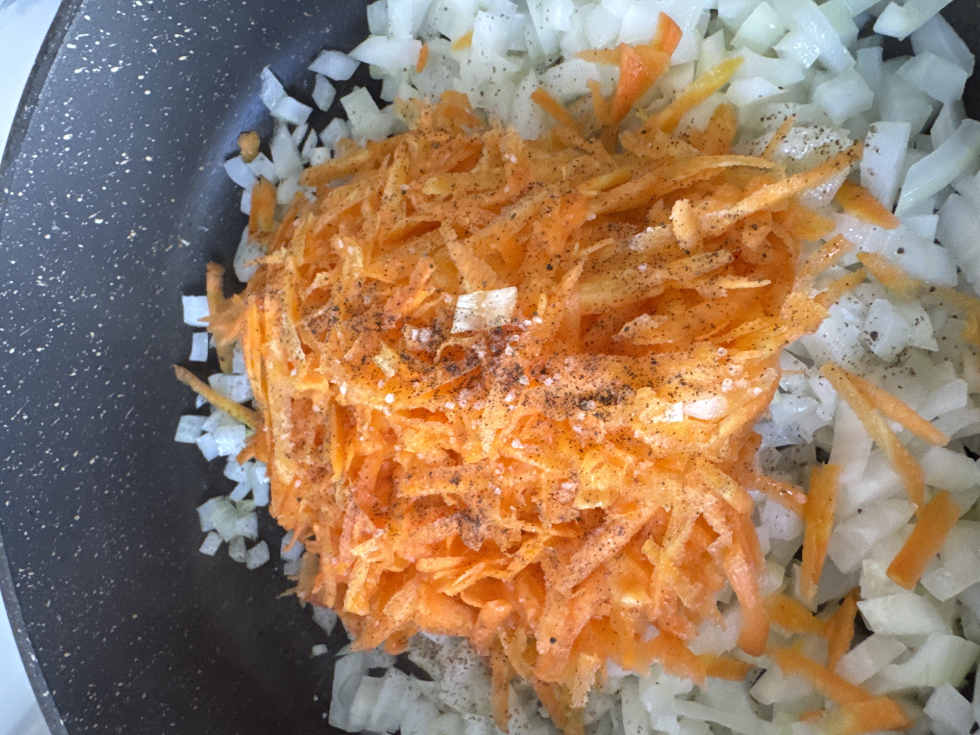 A top-down, close-up shot shows chopped onions and shredded carrots in a dark-colored skillet. The diced onions appear predominantly white, forming a bed around the shredded carrots. The carrots, bright orange, are piled in the center. Black pepper and salt flakes are sprinkled over the carrots and onions. The skillet's surface is speckled. The image does not depict any information that would permit determining a specific location.