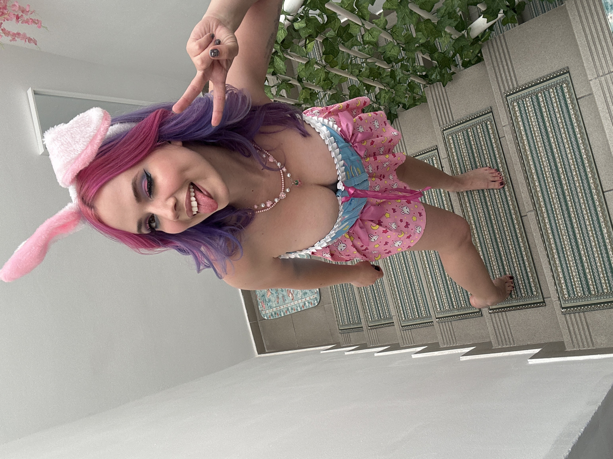 A young woman with dyed pink and purple hair is wearing pink bunny ears and a pink dress with blue trim. She is sticking her tongue out and making a peace sign with her fingers. She is wearing a pearl necklace. She is standing on a tiled floor with a green plant in the background. The location is not discernible from the image.