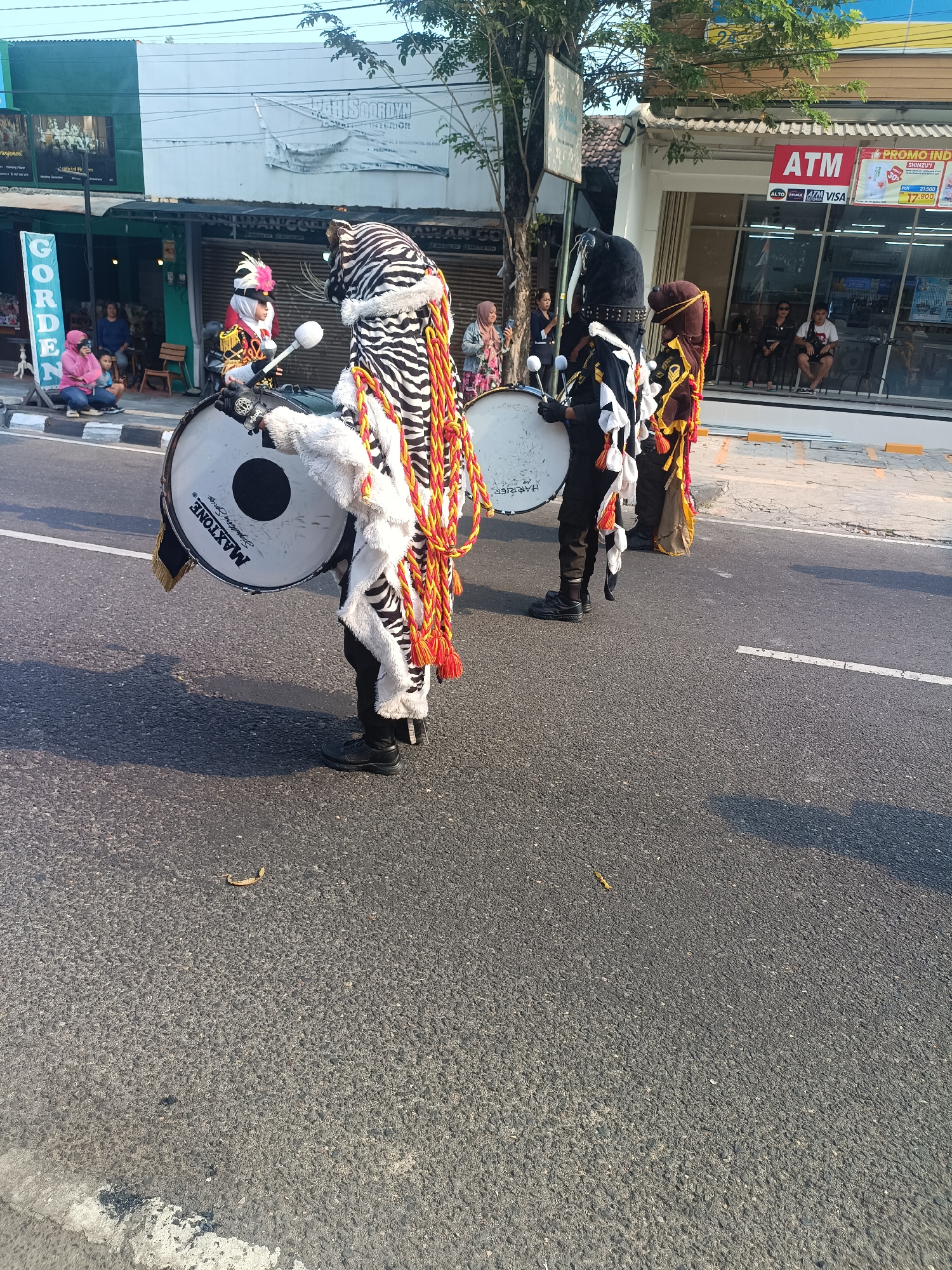 In Sleman, Indonesia, a street parade features individuals in elaborate costumes playing drums. One participant is dressed in a black and white zebra-patterned costume with red and yellow tassels, holding a large drum labeled "MAXTONE". Another individual wears a black helmet with studs and a white fringe, also holding a drum. A third person is clad in a brown headdress and a costume with yellow trim. In the background, spectators are visible along the street, some sitting and others standing. Buildings line the street, including one with an "ATM" sign and a store displaying various products. The event takes place on a paved road with white lane markings.