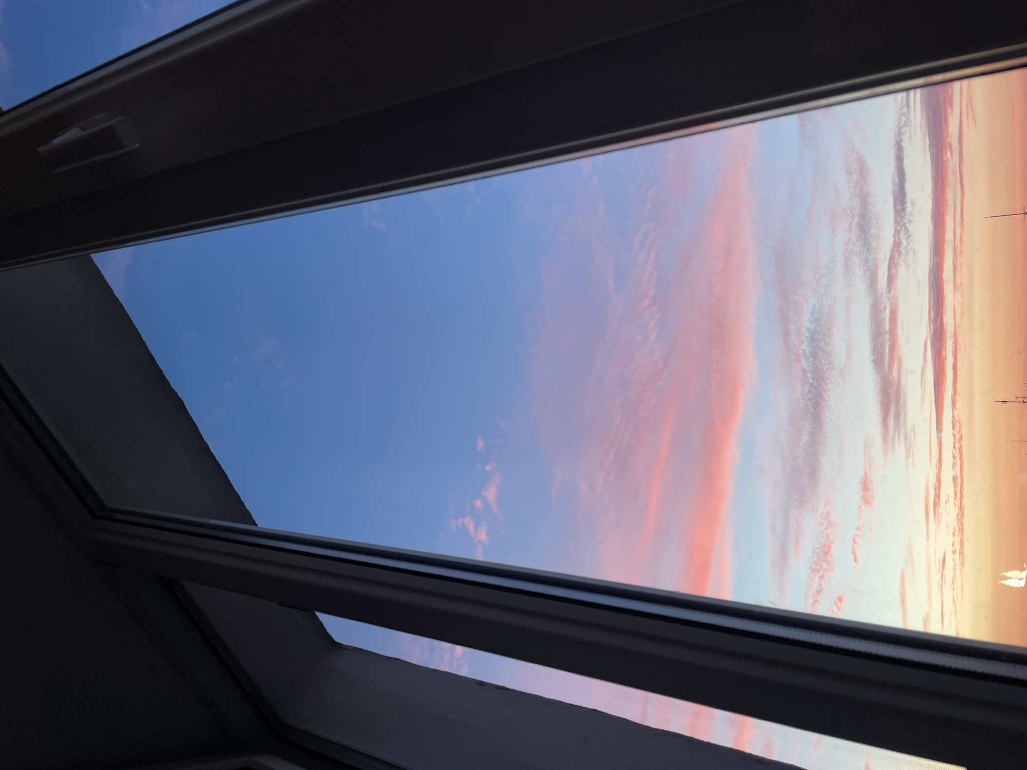 This image shows a view of a sunset sky through a window frame. The sky is a gradient of blue at the top, transitioning to pink and orange near the horizon, with wispy clouds painted in shades of pink. In the distance, silhouetted against the bright horizon, are what appear to be masts of boats and possibly a distant building or structure. The window frame, which is dark in color, occupies the left and right edges of the image, partially obscuring the view. The scene suggests an urban setting, possibly overlooking a body of water, given the presence of boats. The time of day is clearly sunset, with the sky displaying typical sunset colors. Without further visual cues, it is not possible to definitively pinpoint the location within Hrodna, Belarus.