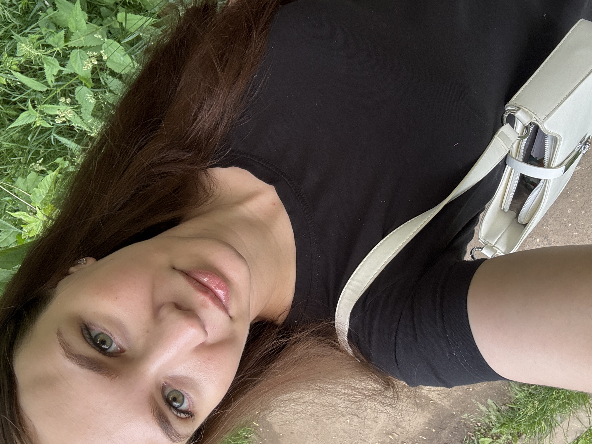 An outdoor, medium shot shows a young woman with long, dark brown hair and green eyes. She is wearing a black t-shirt and a white purse with a silver zipper is slung over her shoulder. She is looking into the camera with a slight smile. The background consists of green grass and some dirt. The location is the city of Chaikovsky, Russia.