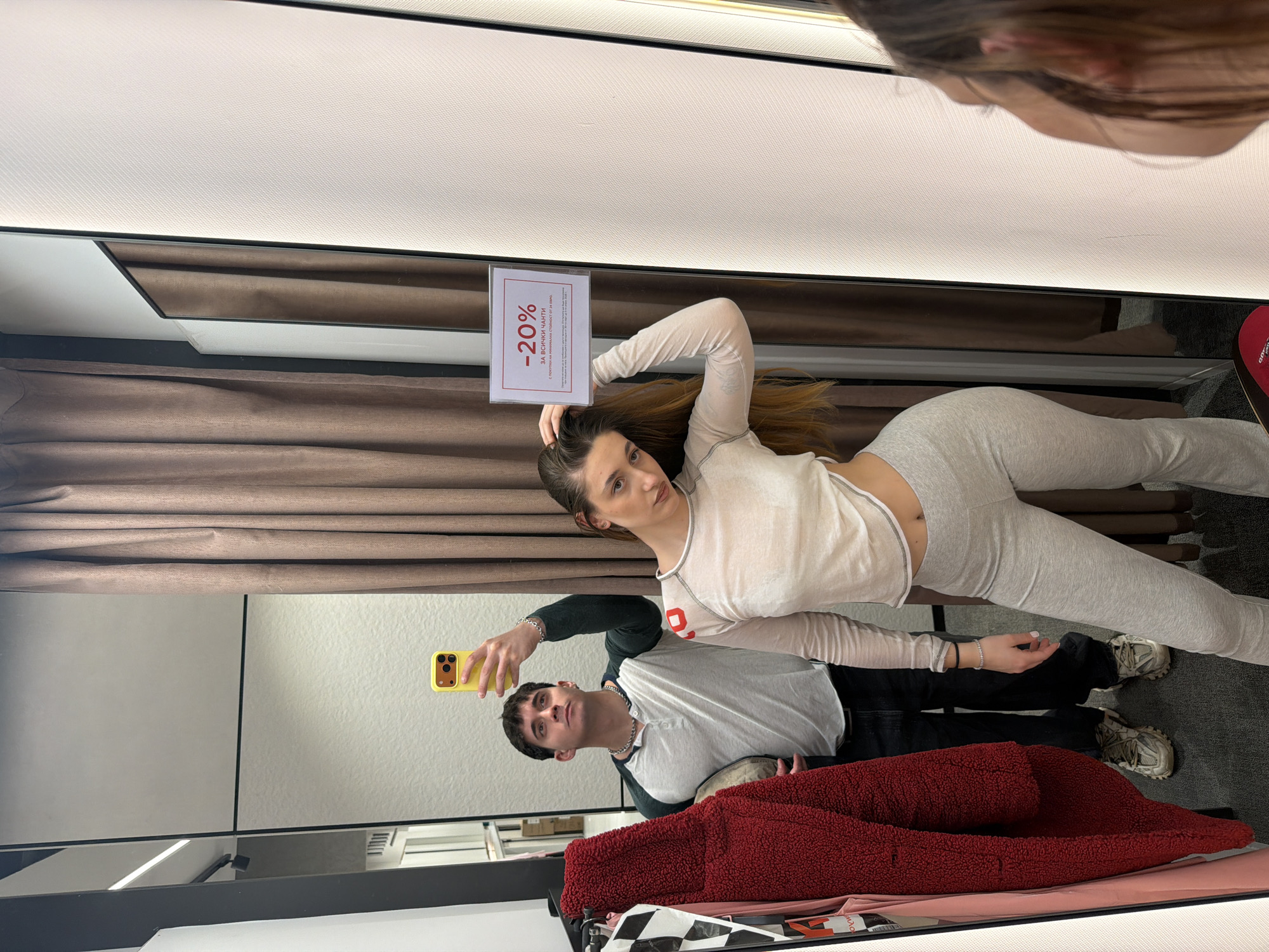The image shows a woman lying on a bench in a changing room of a clothing store in Sofia, Bulgaria. She is wearing light grey lounge wear (long sleeved top and pants) and her hair is light brown. She is holding a sign that says "-20%". A man, possibly her partner, is standing to the side and taking a photo of her with a yellow phone. He is wearing a dark green/black vest over a white short-sleeve shirt, and a silver chain. A red jacket and some clothing items are lying on the bench near them. The changing room has light-colored walls and a brown curtain.
