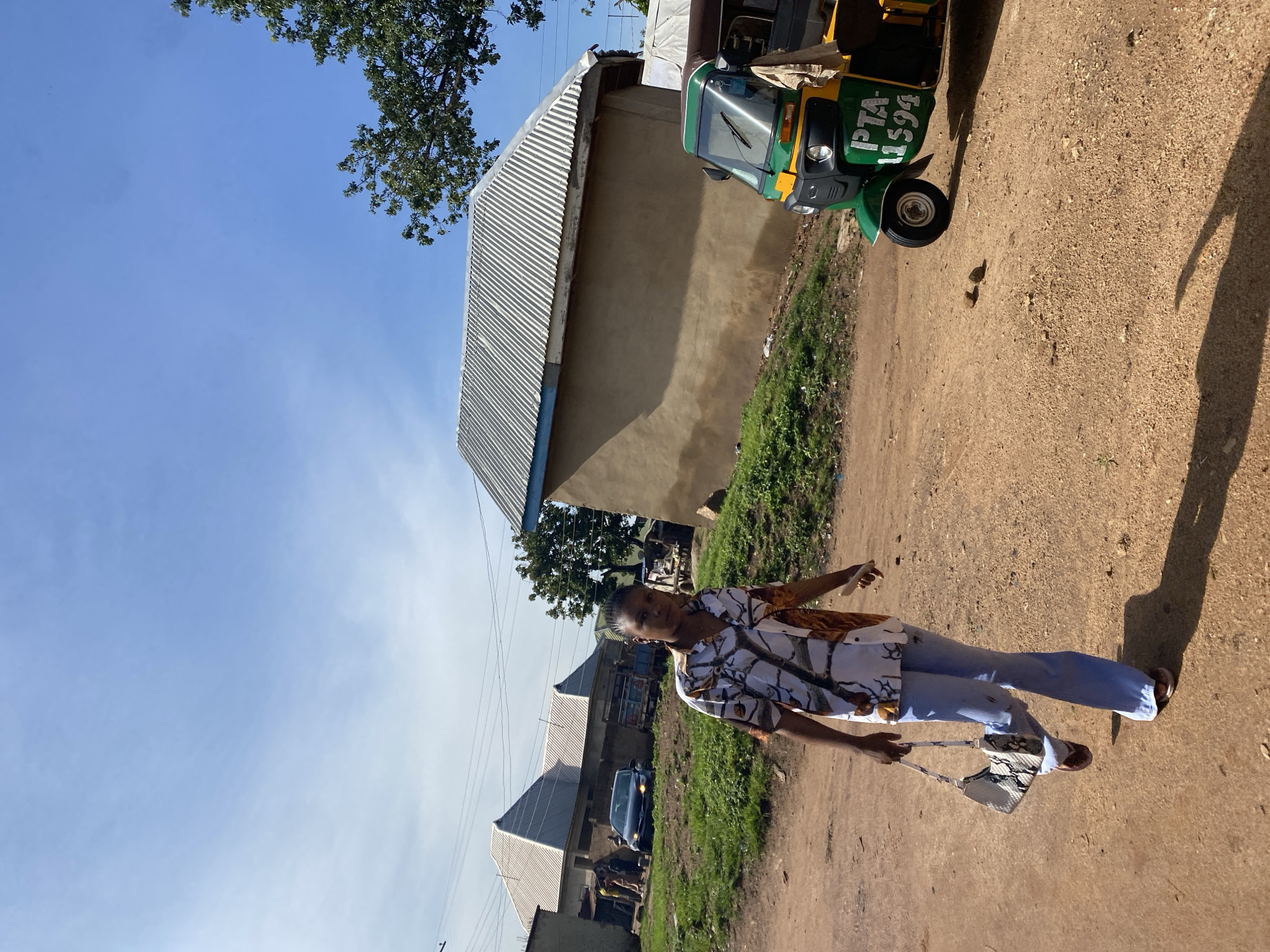 In Barakin Lerawi, Nigeria, a woman stands on a dirt road, facing the camera. She is wearing a patterned short-sleeved shirt and light blue flared pants. A patterned handbag is slung over her left shoulder. To her right, a green and yellow three-wheeled auto-rickshaw, identified by the license plate "PTA 11594", is parked on the side of the road. In the background, buildings with corrugated metal roofs are visible, along with some greenery and trees. The sky is clear and blue. Other people and vehicles are present in the background, suggesting a residential or semi-urban area.