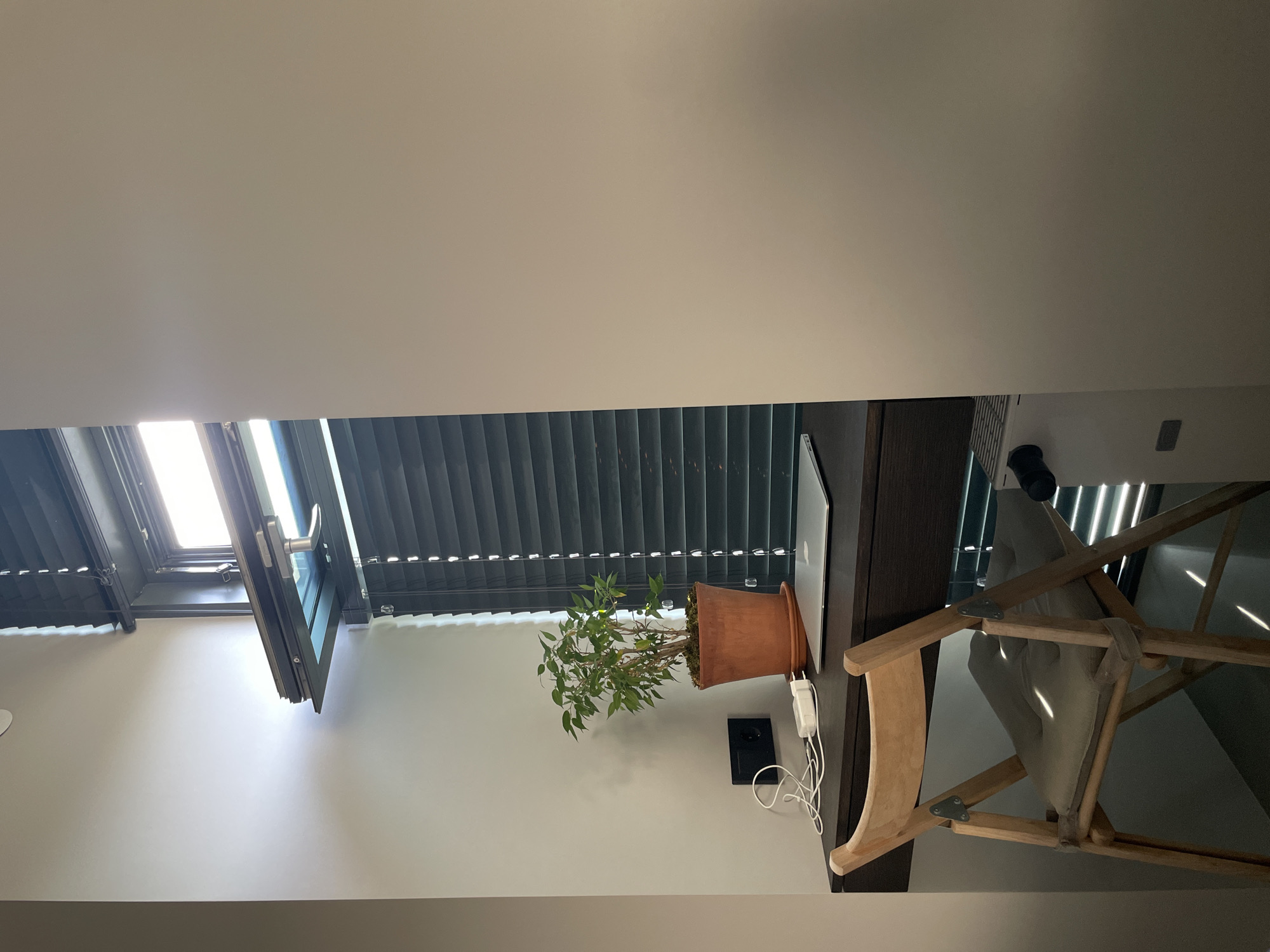 An interior shot depicts a minimalist desk setup against a wall with a window. The desk, made of dark wood, is positioned in front of dark horizontal blinds. On the desk, a laptop sits open, next to a terra cotta pot holding a small green plant. A white phone charger and coiled white wire are also visible on the desk, along with a black electrical outlet on the wall. A wooden folding chair with a light gray cushion is placed in front of the desk. Sunlight streams through the blinds, casting striped shadows. To the right, a portion of a light-colored wall dominates the frame, with a small part of a white appliance visible in the lower right corner. The overall impression is one of a quiet, modern workspace.