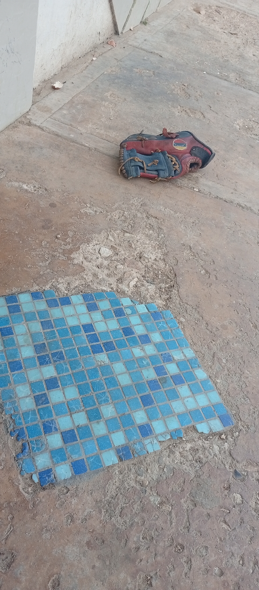 The image captures an outdoor scene, likely during daylight hours, focusing on a red and blue baseball glove resting on a rough, textured ground surface. The ground appears to be a mix of worn concrete and dirt, showing signs of age and possibly neglect, with cracks and debris visible.
In the upper central part of the frame, a baseball glove is positioned. It features dark red or maroon leather on the back and wrist area, with dark blue or grey leather forming the palm and fingers. Light brown lacing is used to hold the glove together. A circular patch with a stylized graphic, possibly a logo or emblem, is visible on the glove's back, though any specific text is illegible.
Towards the lower left, a square section of blue mosaic tiles is embedded in the ground. These tiles vary in shades of light and dark blue, arranged in a grid pattern. Some tiles along the edges appear chipped, broken, or missing, indicating wear and tear. The ground immediately surrounding these tiles is particularly uneven and coarse.
A light-colored wall or base of a structure runs along the top and left edges of the image, forming a boundary for the ground area. No human subjects are present, and no particular activity is depicted. The bright and even lighting suggests a clear, sunny day. The scene appears to be an outdoor area in Juan Griego, Venezuela, possibly a yard, path, or part of an abandoned structure.