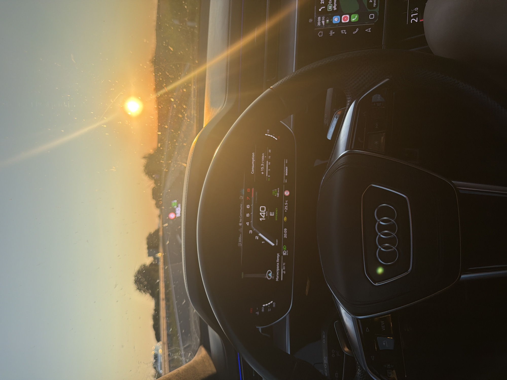 The image is a first-person view from inside a car, likely an Audi, at sunset. The sun is low on the horizon, casting a warm glow and creating a lens flare across the windshield. The dashboard displays are active, showing speed (140 km/h), efficiency information, and the current time (20:09). A navigation map is also visible on the right-hand display. The car is on a highway, with trees and fields visible outside, suggesting a rural or suburban setting. The windshield has water droplets on it. The driver's steering wheel is prominent in the foreground, with the Audi logo visible. The temperature inside the car is indicated as 21.0°C. No people are visible in the image. The location is identified as Gabarnitsa, Bulgaria.