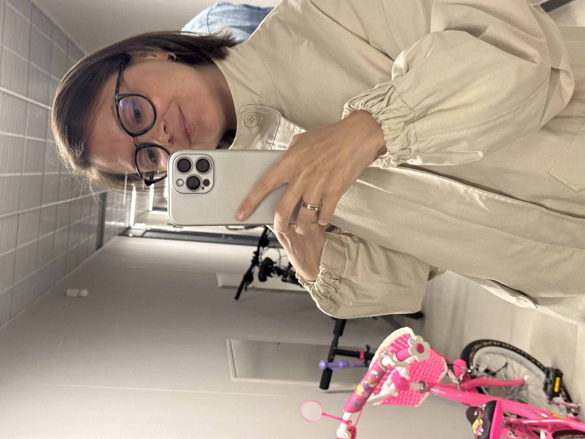 A woman with brown hair and glasses takes a selfie indoors, holding a smartphone. She wears a beige jacket with gathered sleeves. A bicycle, partially visible, sits behind her. The scene appears to be in a hallway or storage area, with tiled walls visible in the background. The presence of the bicycle suggests a residential setting. No specific information indicating the location is Izhevsk, Russia, is evident in the image.