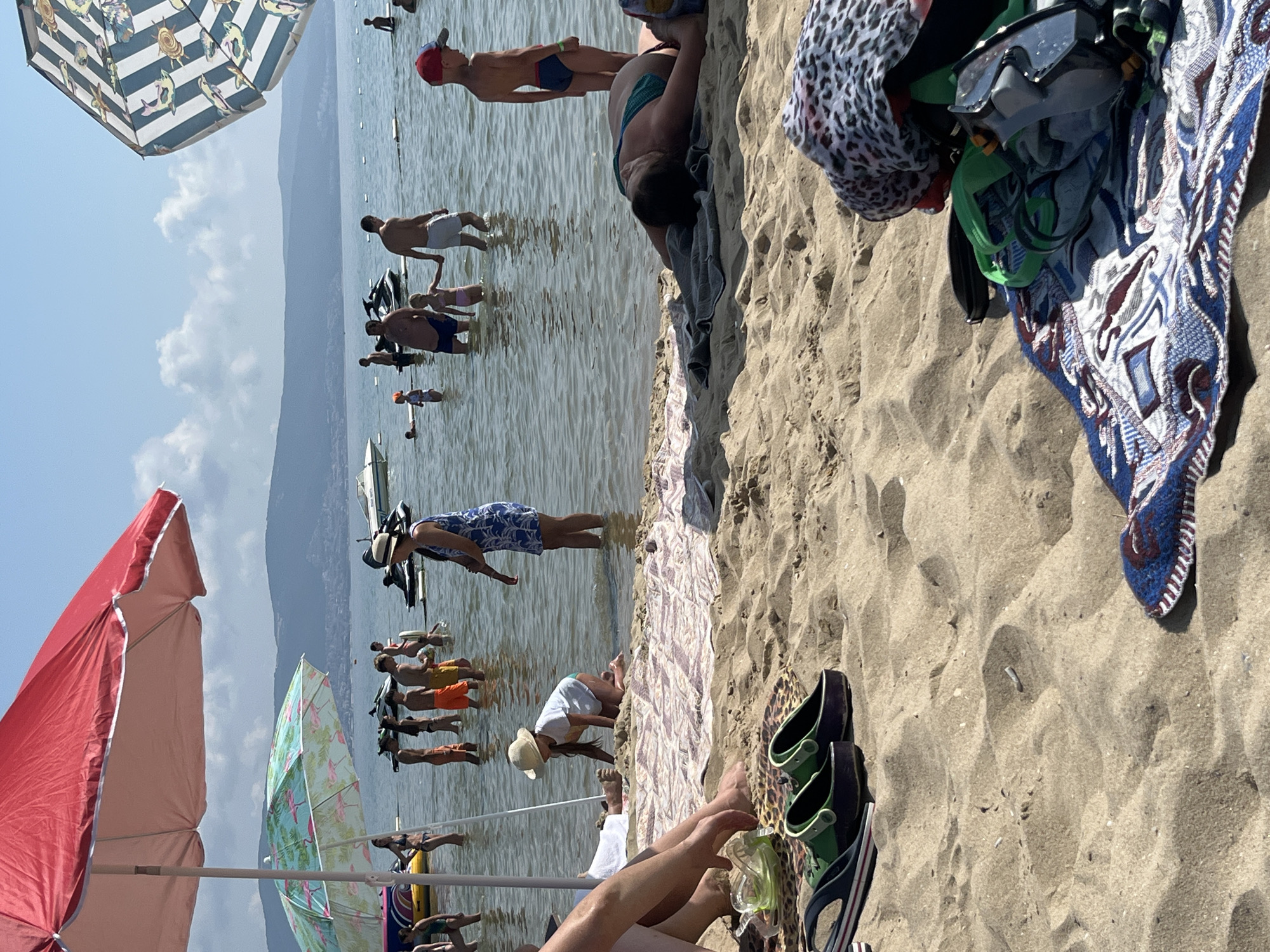 The image depicts a crowded beach scene in Nesebar, Bulgaria, under a clear blue sky with scattered clouds. In the foreground, sand covers the ground with several items of clothing and accessories scattered on it, including two pairs of clogs, a towel with a patterned design, and a leopard-print scarf draped over a pile of belongings including a snorkel mask and goggles. Further into the scene, numerous people are wading in the shallow, clear water of the Black Sea. Some are standing, while others, particularly children, are playing near the shoreline. Several beach umbrellas, one large red one and a smaller one with a flamingo pattern, provide shade. In the background, the sea extends to a distant hazy shoreline with mountains, and several jet skis are visible, indicating recreational activities. The overall atmosphere is one of a lively summer day at a popular tourist destination.