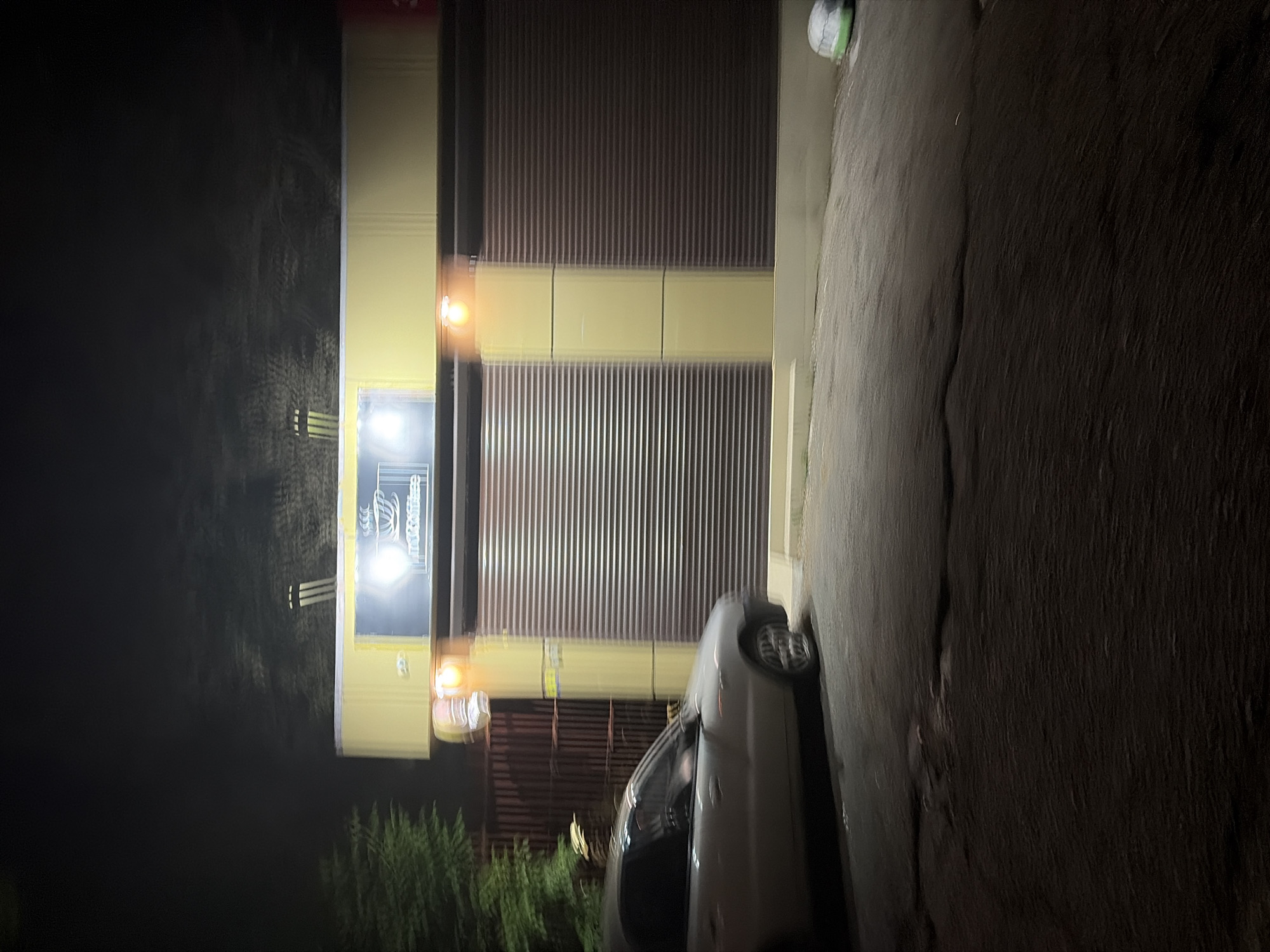 The image, taken at night, displays a light-colored car parked on a paved surface alongside a commercial building. The scene suggests an urban or semi-urban environment, possibly a service or industrial area in Sochi, Russia.
The central subject is a building with a light-colored facade featuring prominent dark, corrugated roll-down shutters. A horizontal band, possibly part of the building's trim or signage support, runs across the facade and is illuminated by several glowing yellow-orange light bulbs. Below this band, to the left, is a brightly lit rectangular sign. The sign clearly displays the word "ЛЕТО" (LETO, meaning Summer) in a stylized font, with "Containers" visible in English text beneath it, likely indicating the business name "Summer Containers" or a similar storage/logistics service.
A light-colored (possibly silver or white) sedan or hatchback car is parked parallel to the building on the right side of the frame. Only its rear and side profiles are visible, with its wheels turned slightly towards the building.
The foreground and the area beneath the car consist of a dark, paved surface, possibly asphalt or concrete, which shows some texture and minor cracks. To the far left, in the dark background beyond the building, some indistinct, dark vegetation (likely trees or bushes) can be faintly discerned. The sky is completely dark, confirming it is nighttime. The only sources of light are the artificial illuminations from the building's sign and bulbs. No human figures are visible, and the scene is static.