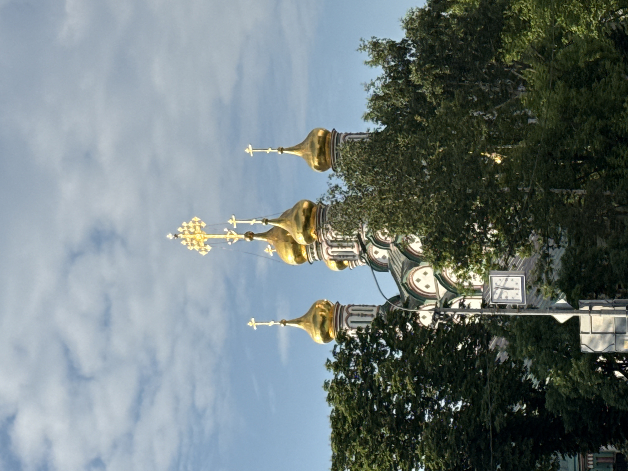A low-angle, full-shot photograph captures the gilded domes of the Church of the Transfiguration of the Savior in the Ostankino district of Moscow, Russia. The church's domes, topped with crosses, emerge from behind a dense canopy of lush green trees. A lamppost with a square clock face displaying the time as approximately 12:35 is visible in the foreground, to the left of the church. The sky is partly cloudy, with patches of blue visible between the white clouds. The overall lighting suggests a bright, sunny day, possibly late morning or early afternoon, with the sun illuminating the golden domes and casting shadows on the trees. The architectural style of the church, with its multi-tiered domes and intricate detailing, is characteristic of Russian Orthodox churches. The image is framed to emphasize the grandeur of the religious structure against the natural backdrop of the trees and the sky.