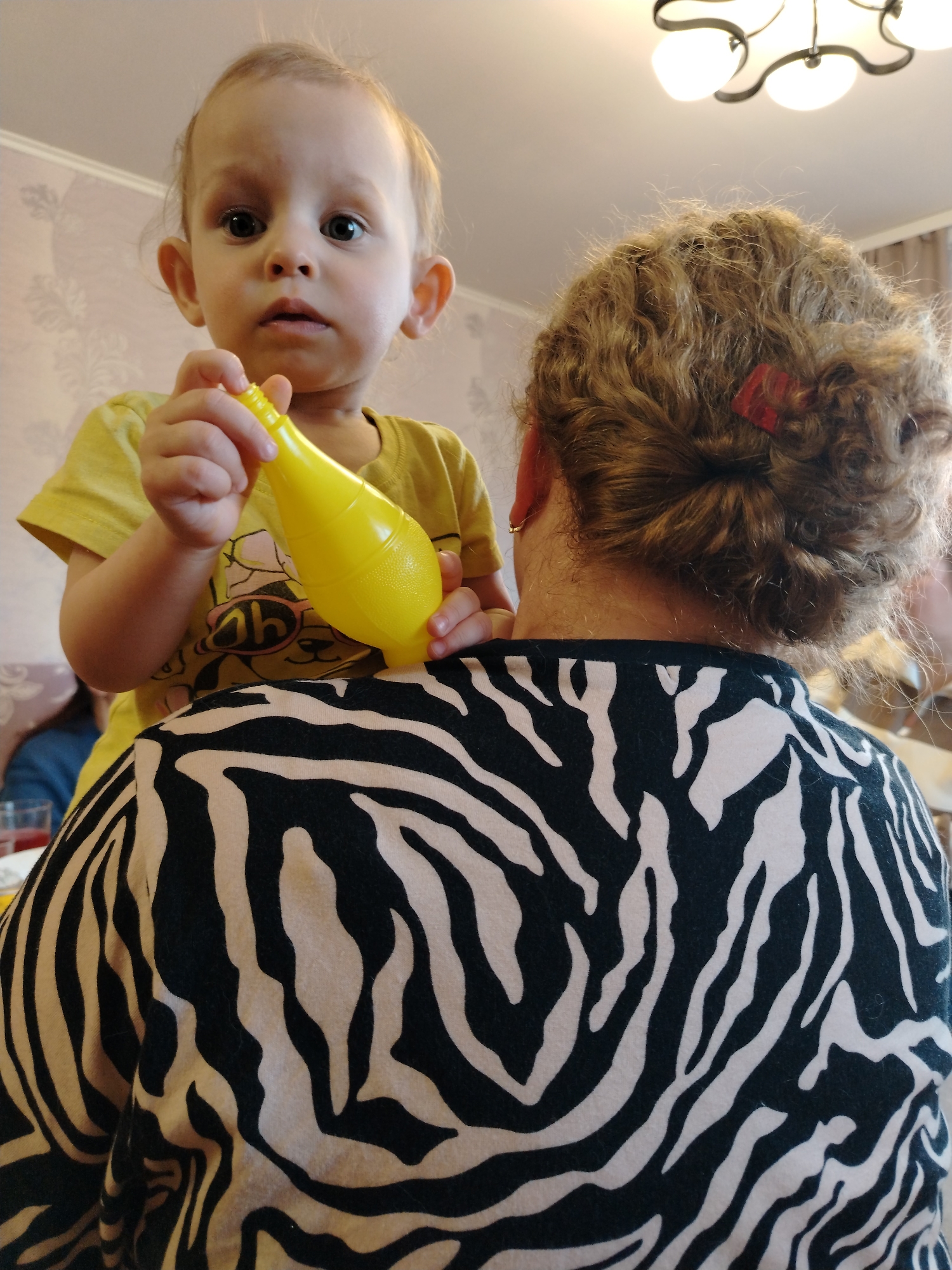 A toddler is sitting on the shoulders of a woman in an indoor setting. The toddler is wearing a yellow shirt and holding a yellow plastic object. The woman is wearing a black and white zebra-striped shirt. A chandelier is visible in the background. The location is Unknown, Unknown.