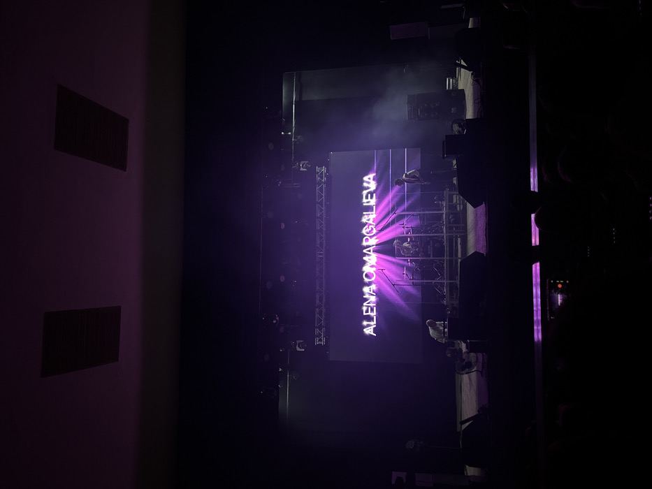 A concert stage glows with violet light. The name "ALENA OMARGALIEVA" is displayed prominently on a central screen, illuminated with bright rays. Stage rigging is visible above. Musicians are dimly lit on risers behind the screen. A dark hall extends on either side, with vents visible on a side wall. The view is taken from the audience perspective.