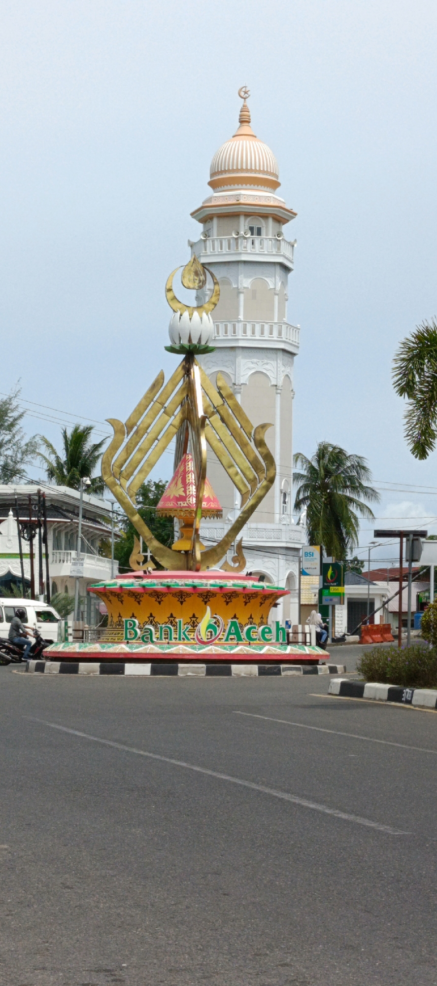 The image depicts the Bank Aceh monument in Banda Aceh, Indonesia, a city in the province of Aceh. The monument is located at a street intersection, with a mosque minaret visible in the background. The monument itself is elaborately decorated, featuring a large yellow bowl-shaped base with intricate patterns. The words "Bank Aceh" are prominently displayed in green lettering, with a stylized logo incorporating a flame-like design. Above the base, a golden, abstract sculpture rises, topped with a white lotus-like element and a crescent moon. Palm trees and other vegetation surround the monument, indicating a tropical climate. Vehicles, including a motorcycle and a white van, are present on the road, suggesting moderate traffic. The sky is overcast. The scene captures a blend of cultural, religious, and commercial elements typical of an urban environment in Indonesia.