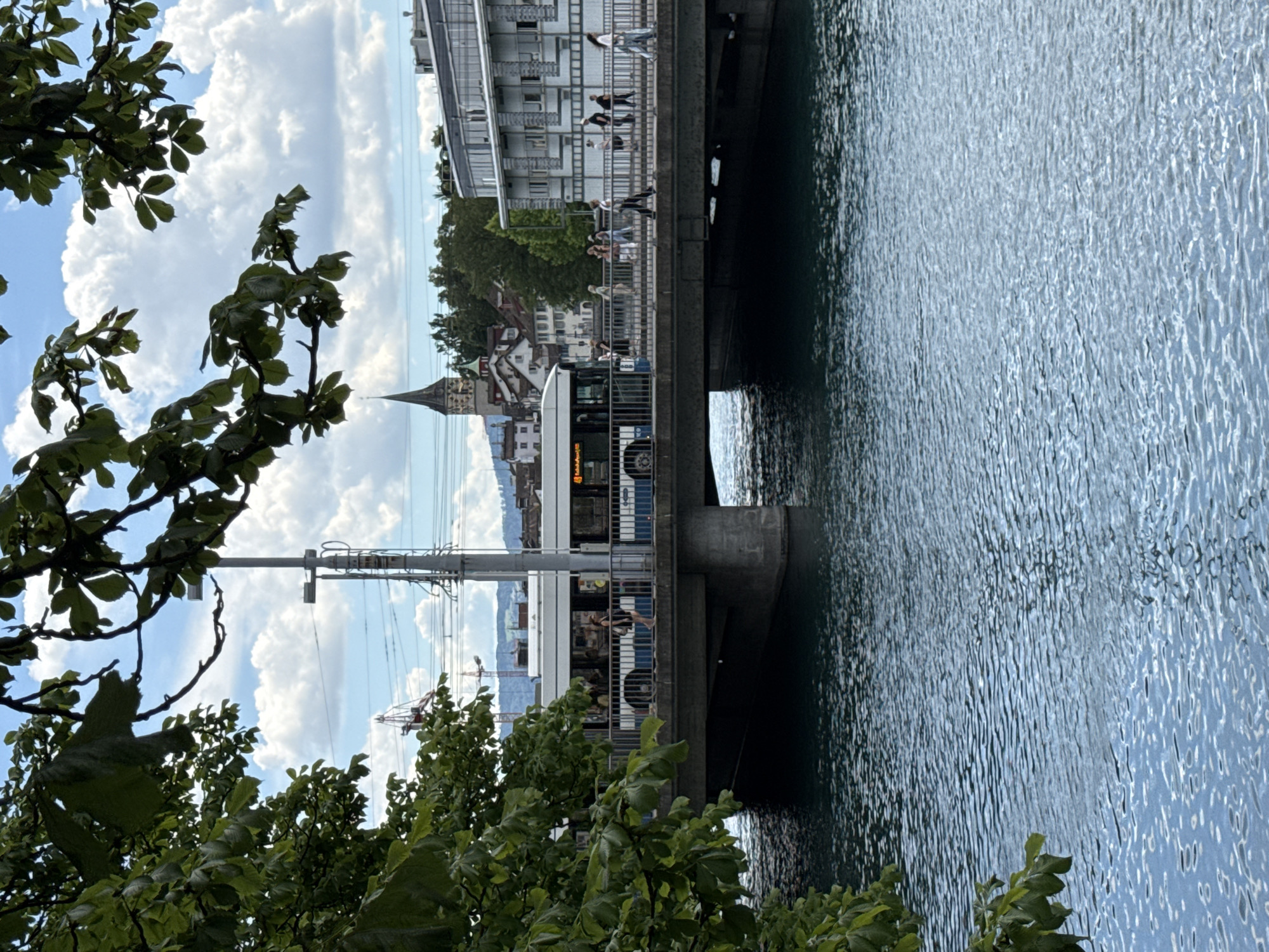 In Zurich, Switzerland, a bus crosses a concrete bridge over a body of water. The bus is white with blue accents and has people inside and on its side windows. On the bridge's sidewalk, pedestrians walk in various directions. In the background, there is a church with a tall, pointed spire, and other buildings with red roofs. Trees with green leaves frame the top of the image. The sky is blue with white clouds. The water is a deep blue-green with ripples.