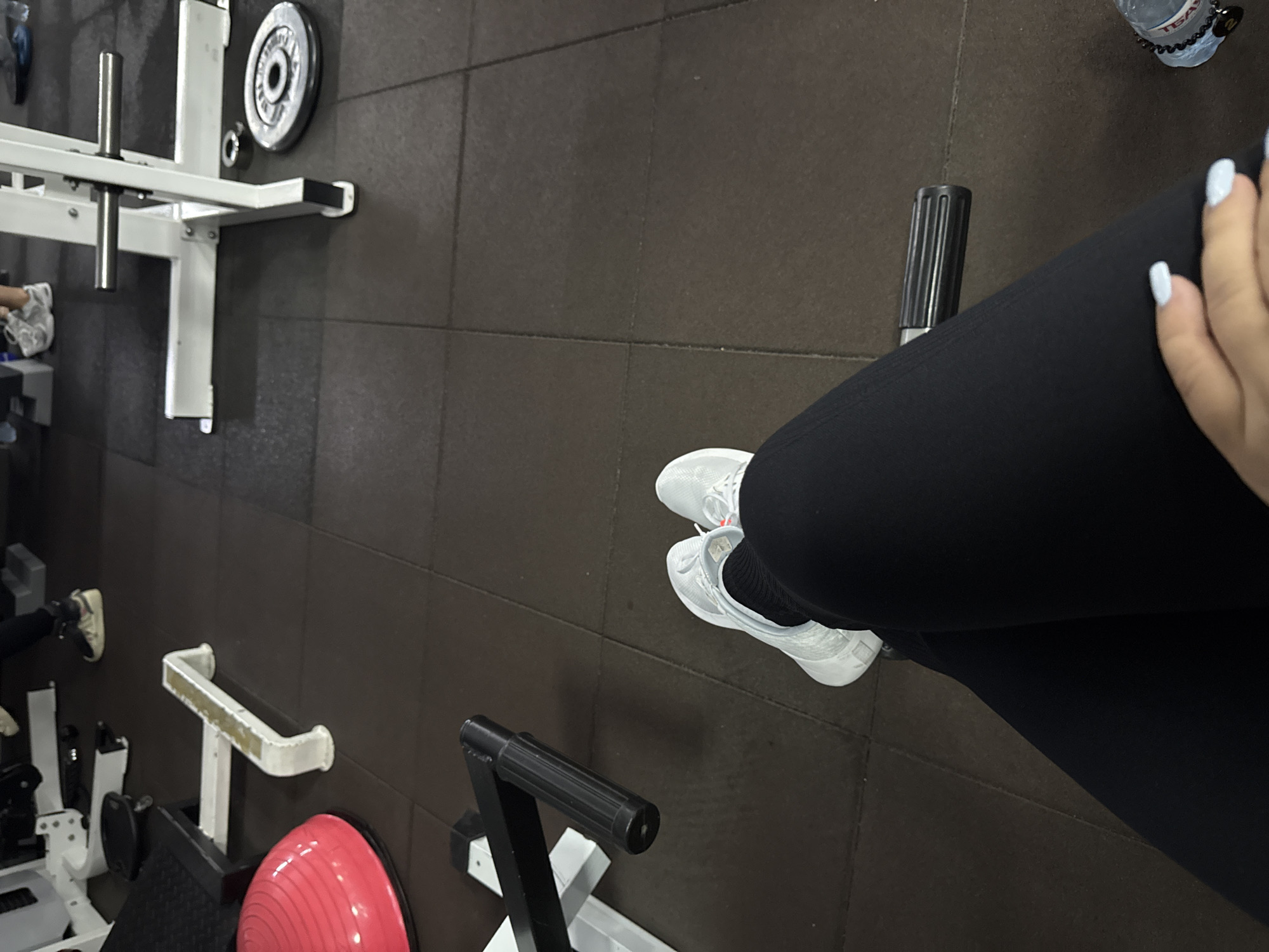 This is a medium-angle, high-point-of-view, indoor shot of a gym in Nazran, Russia. The image is dominated by dark brown rubber flooring with visible seams. In the foreground, a person's legs, clad in black leggings and white athletic shoes, are bent at the knees and resting. The person's right hand, with light blue painted fingernails, is visible in the lower right corner. To the left of the person's legs, a black textured handle of a gym machine is prominent. Further into the background, on the left, a bright red exercise dome is partially visible. A white piece of gym equipment with a bar is positioned in the mid-ground, and a weight plate is on the floor to the right of it. Other gym equipment, including a squat rack and weight plates, are visible in the blurred background, suggesting an active workout environment. The lighting is artificial and casts shadows across the floor.