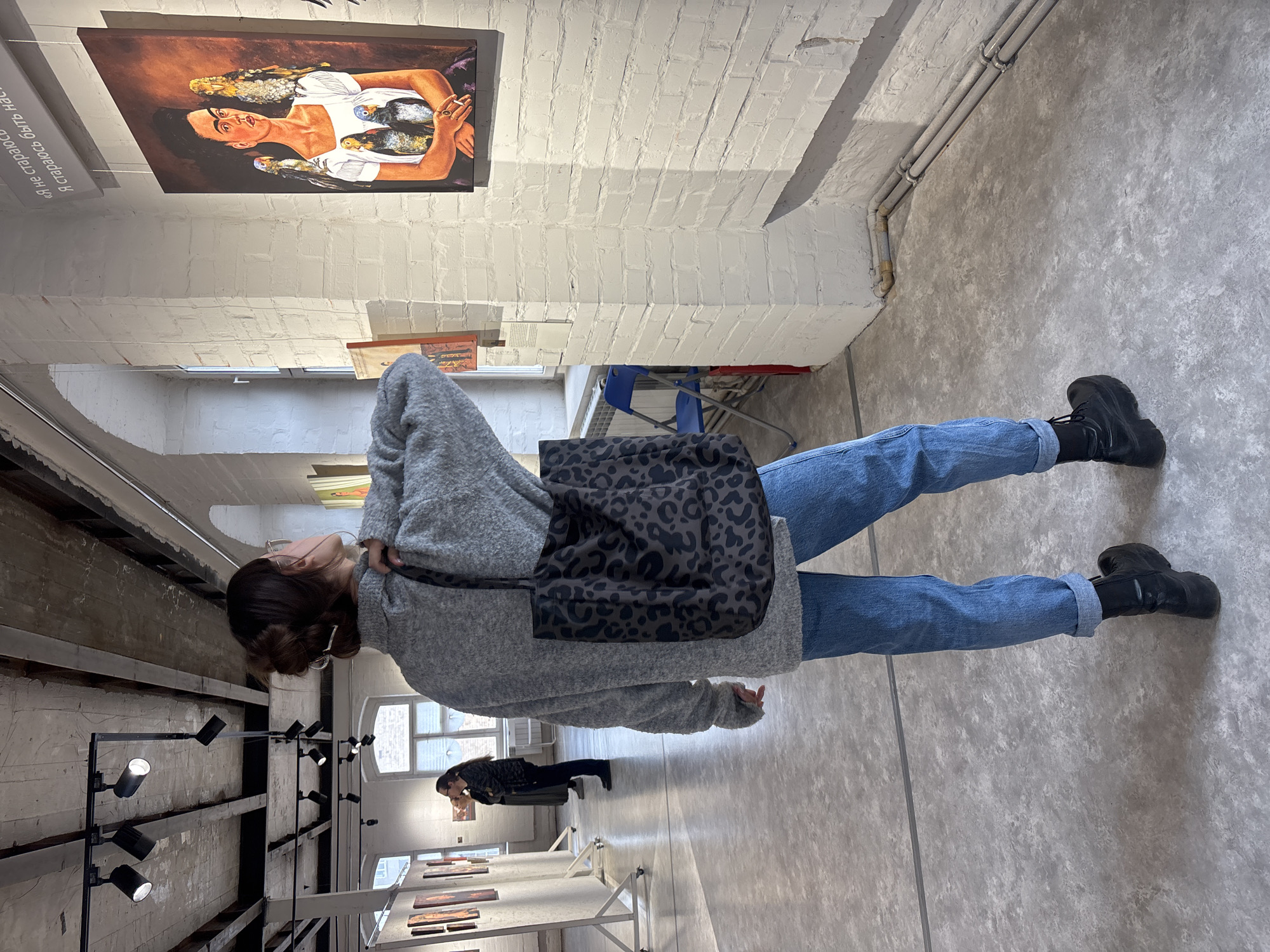 A woman in jeans and a gray sweater stands in an art gallery in Krasnoyarsk, Russia. She wears black boots and carries a black bag with a dark gray animal print. She looks at a painting on the wall, which depicts a woman in a white dress with birds around her. Other art pieces are displayed on the walls and on stands in the background. Another person is visible in the distance, also looking at the artwork. The gallery has white brick walls, concrete floors, and a metal lighting system.