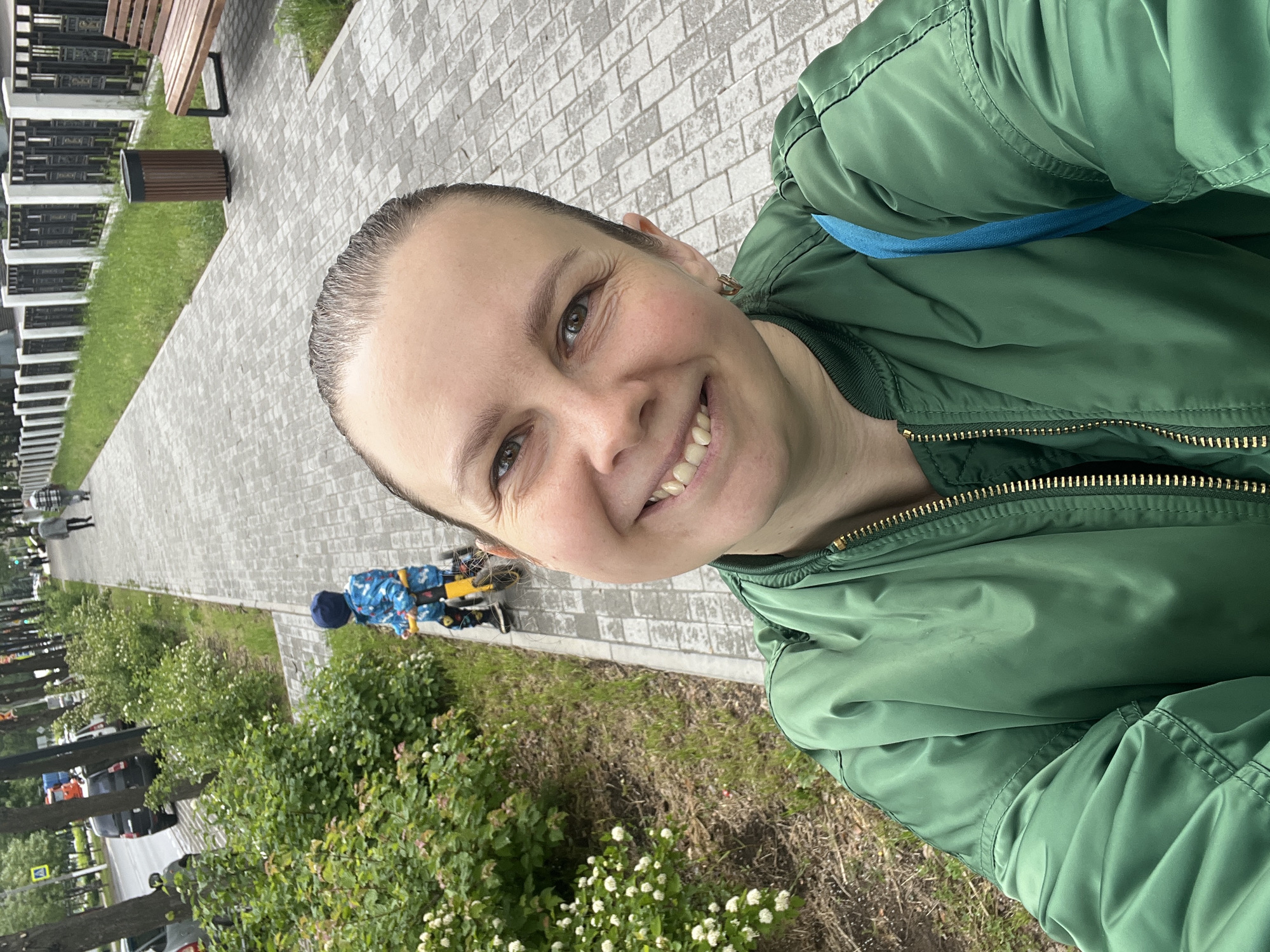 A smiling woman takes a selfie outdoors in Chaikovsky, Russia, during what appears to be a pleasant daytime. She occupies the right side of the frame, looking directly at the camera with a wide, cheerful smile that shows her teeth. She has light brown eyes, fair skin, and dark hair pulled back. She is wearing a dark green bomber jacket, with a glimpse of a light blue strap or lining visible on her shoulder.
The background shows a paved pedestrian path made of light grey interlocking pavers, oriented diagonally from the upper left to the lower right. To the woman's left, a grassy strip is lined with various green bushes, some bearing small white flowers. Further down the path, a small child, dressed in a blue and white patterned outfit and a blue hat, is visible riding a yellow balance bike. In the distance, other blurry figures are walking.
Beyond the path and green spaces, a street with parked cars and some trees are visible. Buildings with black and white railings can be seen in the far background. Near the top left of the image, a wooden park bench and a brown cylindrical trash can are situated beside the paved walkway. The lighting is soft, suggesting an overcast day or diffused sunlight. The woman's happy expression conveys a sense of contentment. No text is explicitly visible.