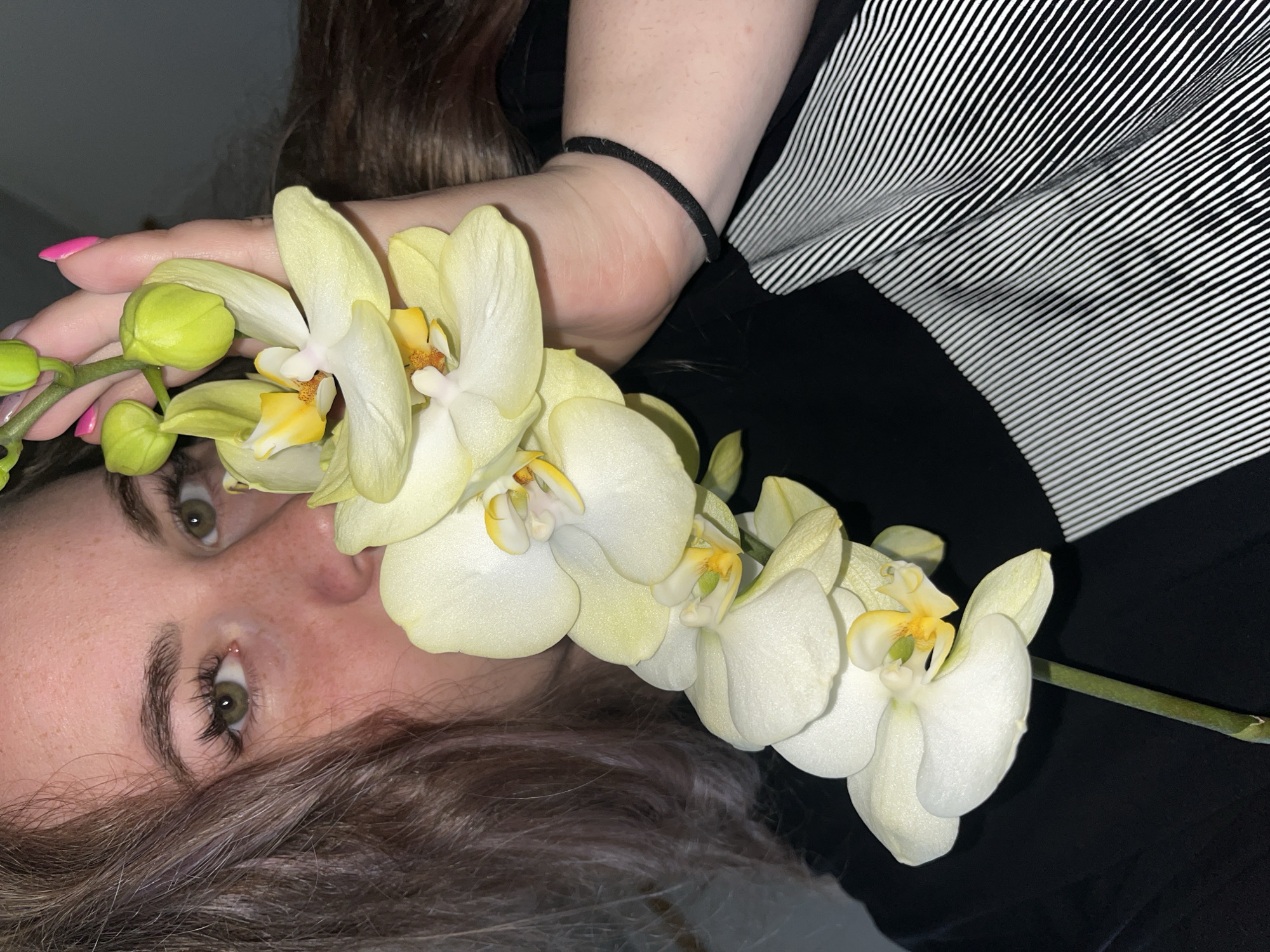 A woman with fair skin and green eyes is holding a stem of yellow and white orchids. Her hair is brown with some grey strands. She has a black elastic band on her wrist. She is wearing a black shirt with black and white striped sleeves. The location is not discernible from the image.