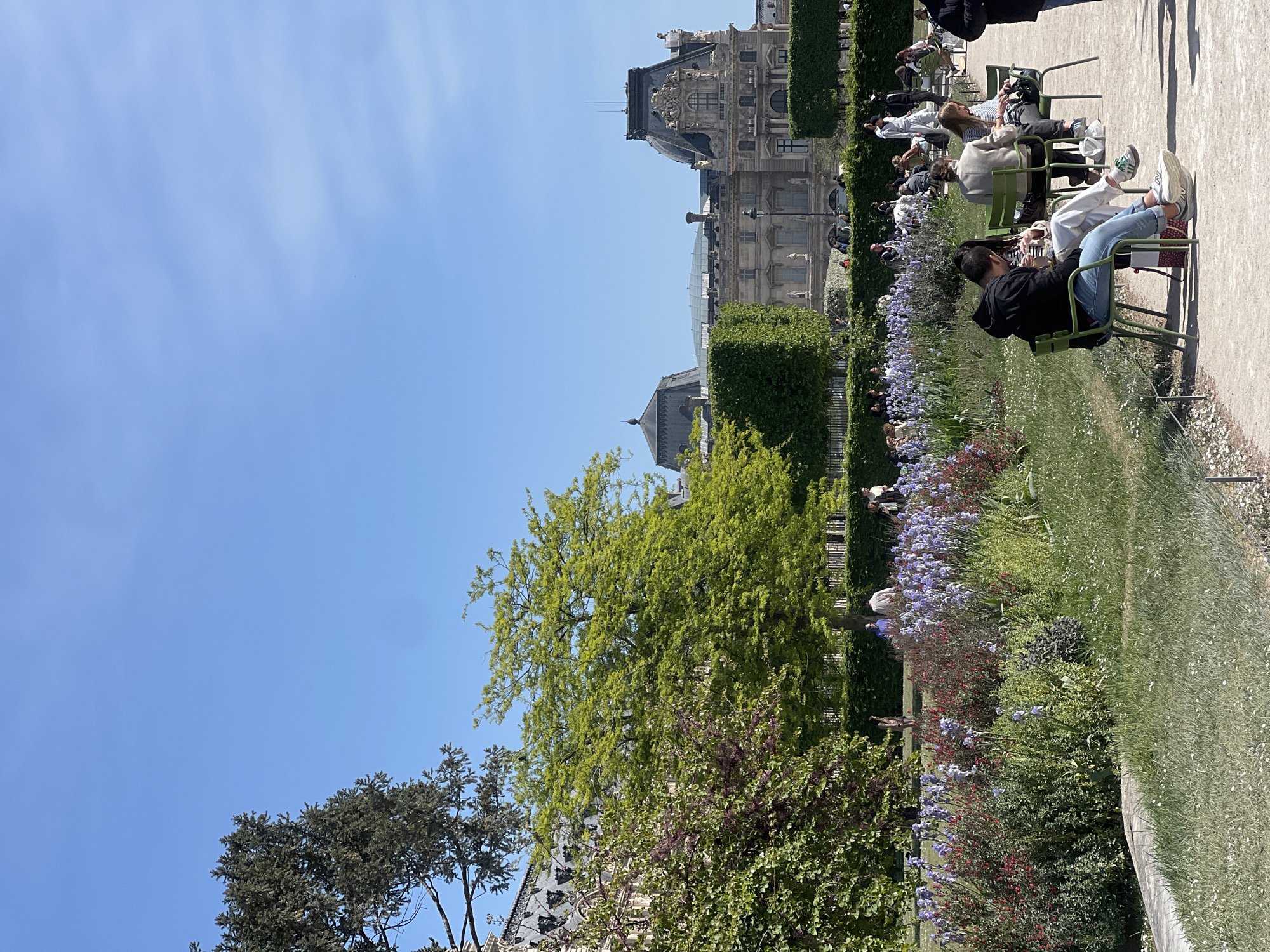 The image presents a serene outdoor scene, strongly indicative of a Parisian garden like the Tuileries, set against the backdrop of a grand historical building. The weather is bright and sunny, with a clear blue sky suggesting a pleasant day, likely during late morning or early afternoon.
In the foreground and midground, several individuals are seen relaxing in iconic green metal garden chairs, positioned on a light-colored gravel path. One person in the immediate foreground, viewed from behind, wears a black top and blue jeans, seated calmly. Beside them, another individual has their legs comfortably propped on a separate chair, highlighting a casual, leisurely atmosphere. Further back, additional people are scattered, enjoying the tranquil surroundings.
The landscape is rich with diverse vegetation. Meticulously maintained hedges create defined borders within the garden. Vibrant flowerbeds burst with purple-blue and red blooms, adding splashes of color. Various trees are visible, including one in the mid-left with bright green, possibly new, spring foliage, and others featuring darker, denser leaves. A well-kept grassy area extends alongside the path and seating.
Dominating the background is a magnificent stone edifice, characterized by classical architecture, intricate sculptures, a distinctive dark slate roof adorned with dormer windows, and tall chimneys. This elaborate structure strongly resembles a wing of the Louvre Palace, situated adjacent to the public gardens. Other less distinct buildings and more greenery can be seen beyond it.
The overall activity depicted is one of leisure and relaxation, with visitors enjoying the pleasant weather and the picturesque setting of a renowned Parisian landmark. No visible text is discernible within the image.