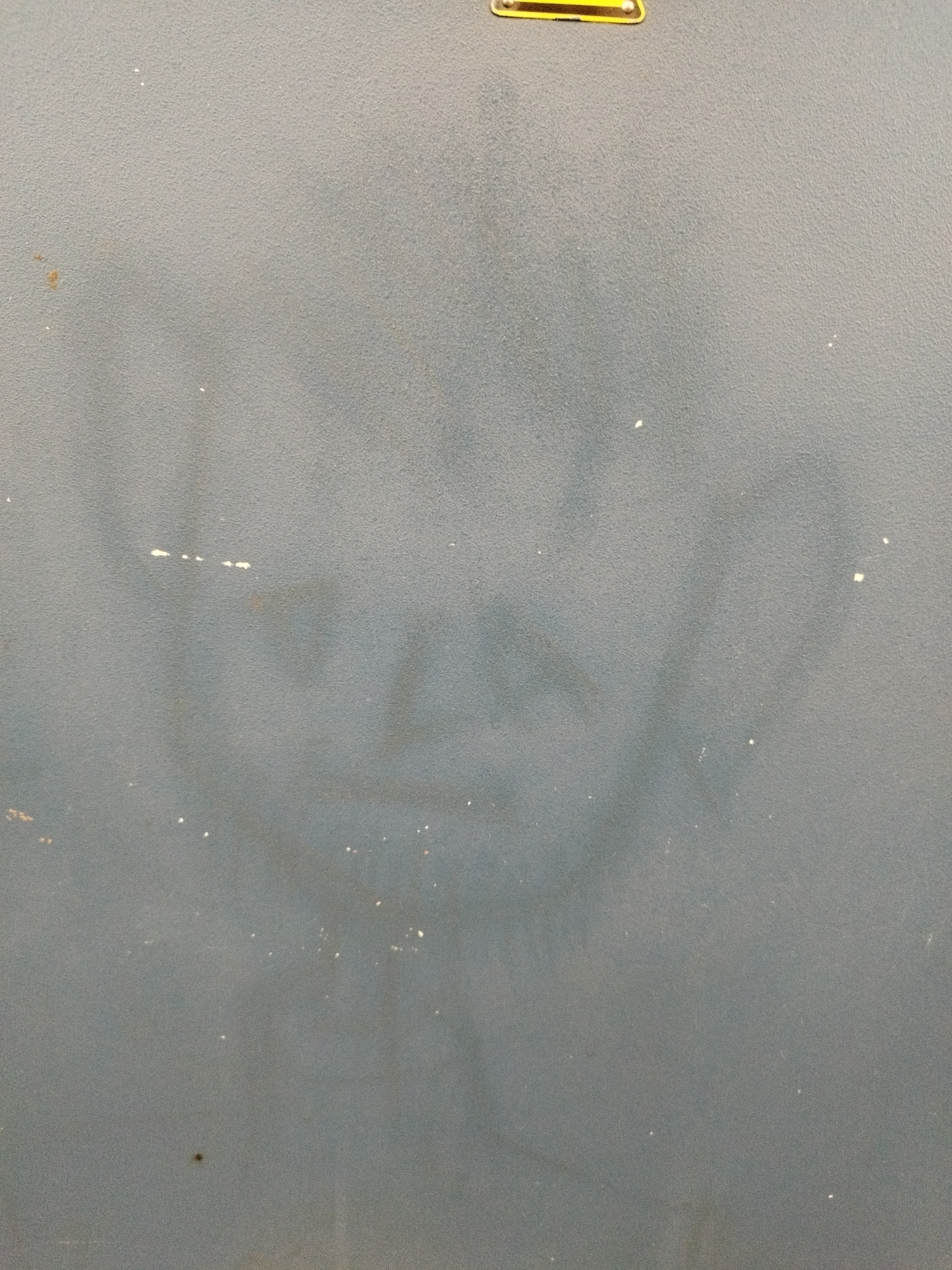 The image shows a rough, gray surface, possibly a wall or a cabinet. There is a faint, smudged image of a face or figure visible on the surface, appearing to be a shadow or a stain. The texture of the surface is granular. At the top of the image, a yellow and black label with some text is partially visible. The image appears to be an interior shot, given the lighting and lack of outdoor elements. There are no people or animals present in the frame. The exact location within Bremerhaven, Germany cannot be determined from the image.
