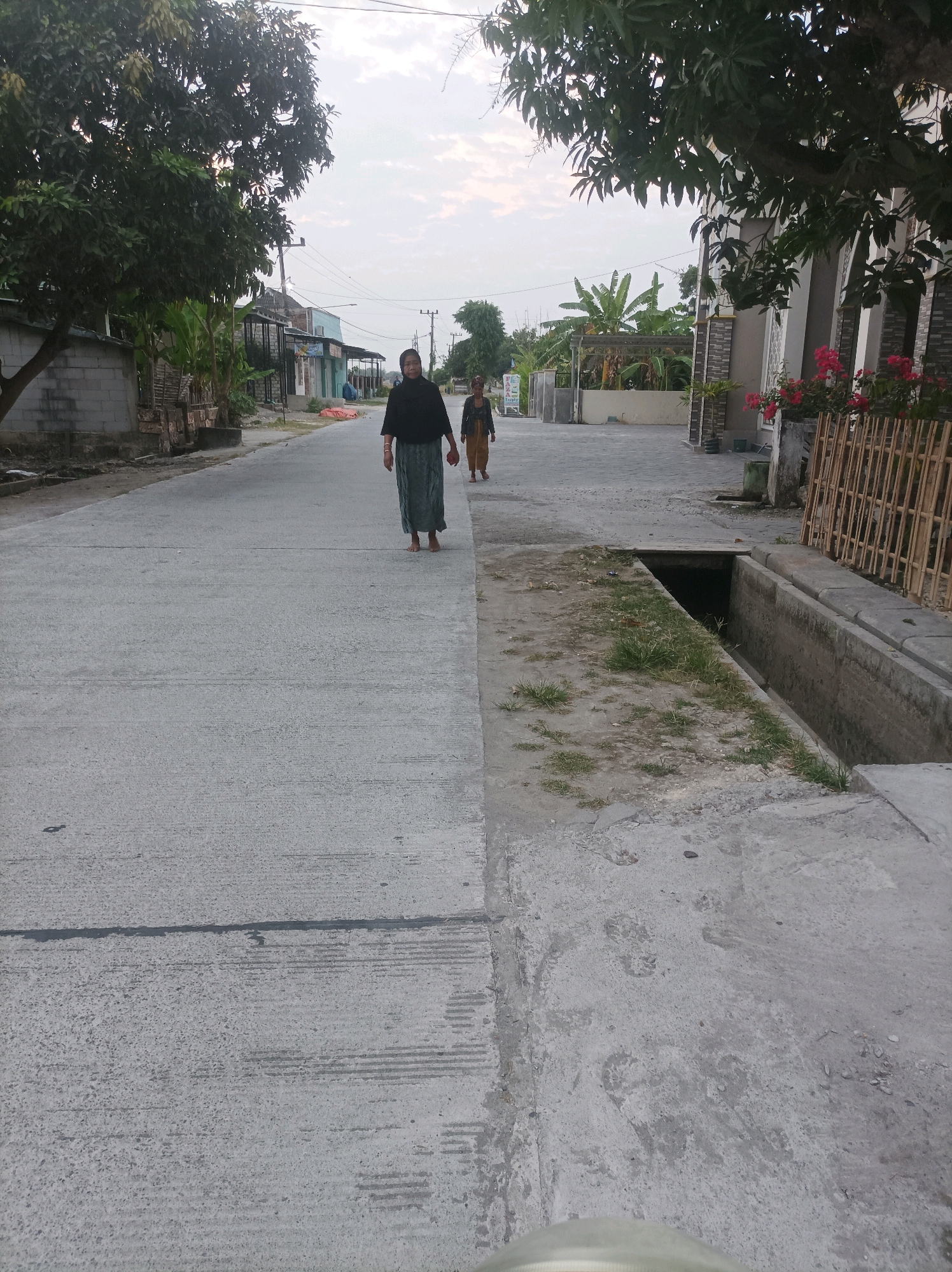 A side street in Canga'an, Indonesia, features two women walking on a concrete road. The woman in the foreground, wearing a dark top and a long, flowing green skirt, walks barefoot. Behind her, another woman in a light brown outfit walks. The street is lined with buildings and vegetation, including trees and banana plants. A concrete drainage ditch runs alongside the road. The sky is overcast.