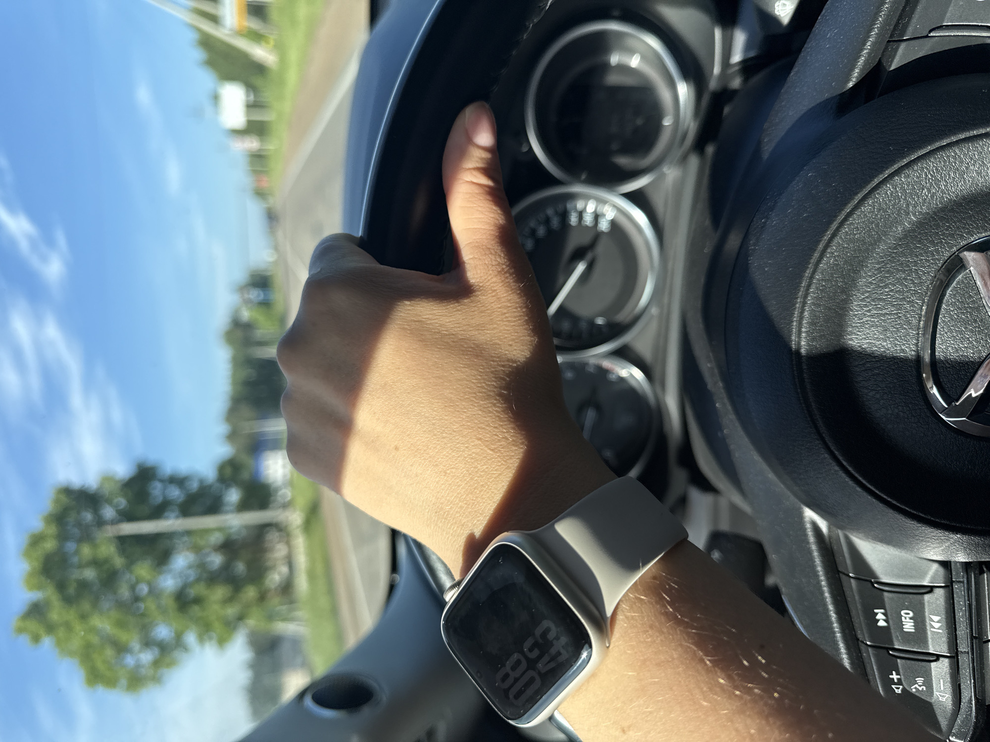 The image captures a first-person perspective from inside a car, showing a person's right hand gripping the top of a black, textured steering wheel. The hand has a light skin tone, and a silver-colored smartwatch with a light gray band is worn on the wrist. The smartwatch screen is visible, displaying digital text: "08" (likely the date or day) and "08:43" (time).
Through the windshield, a bright blue sky with scattered clouds is visible, suggesting clear and sunny weather. Green trees and a blurred road can be seen in the background, indicating the car is in motion. Direct sunlight casts shadows on the hand and parts of the car's interior.
Inside the car, a portion of the dashboard with analogue gauges is visible beyond the steering wheel. To the right of the steering wheel hub, car controls are present, featuring buttons with text and icons: "INFO", track skip arrows ("<4", "4>"), volume controls ("+", "-"), and a speaker/mute icon. A metallic, partially visible emblem, resembling the Mazda logo, is centered on the steering wheel.
The scene depicts the activity of driving a car during the daytime. The location context provided is Ольховка, Russia.