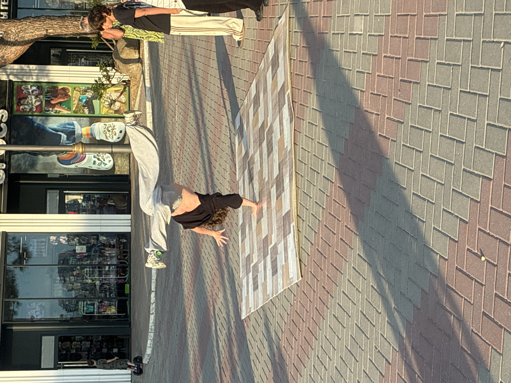 An outdoor shot captures a person breakdancing on a rectangular mat laid on a cobblestone pavement in front of a store. The store appears to be a Crocs shoe shop based on the branding and window displays. The breakdancer, wearing a black top and grey sweatpants, is in the middle of a handstand with their legs extended upwards. The mat has a patterned design of beige, grey, and white rectangular tiles. In the background, a few other individuals are visible, including a young person on a self-balancing scooter to the left and two adults standing to the right. The sunlight casts long shadows across the pavement. The scene is set in Gelendzhik, Russia.