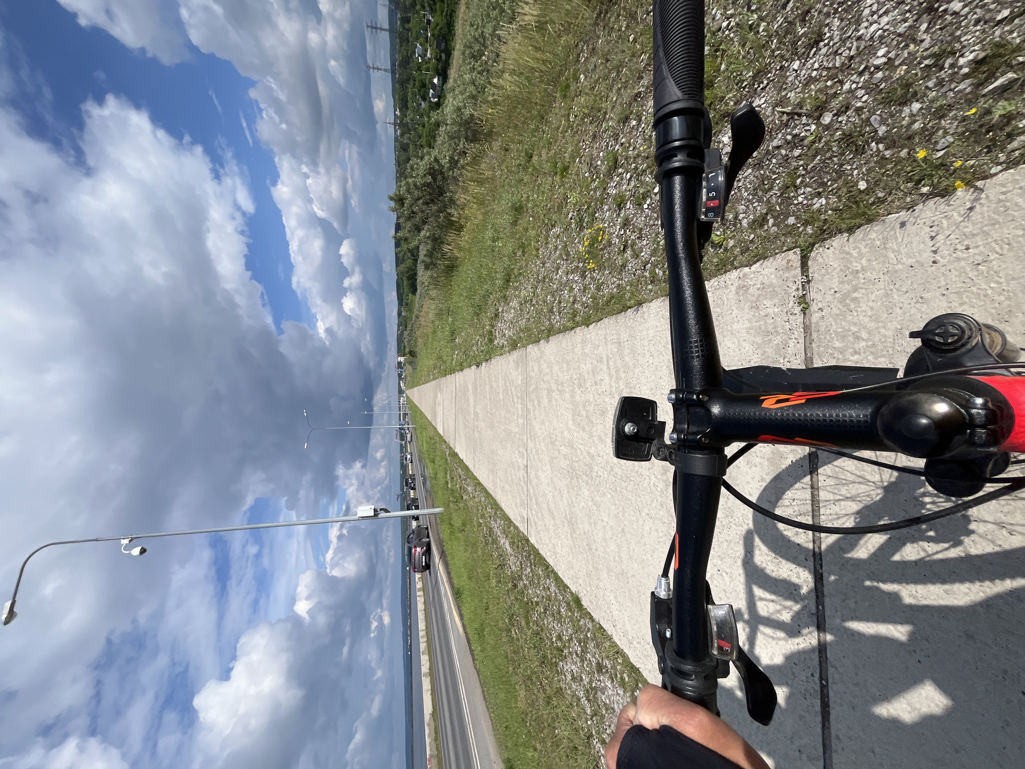 The image captures a first-person perspective of someone cycling on a concrete path next to a road. The road has multiple lanes and traffic, including cars. On the left side of the road is a body of water, possibly a reservoir or river, and beyond that, a shoreline with some buildings. The path is bordered by a grassy embankment with some shrubs and trees. The sky is partly cloudy with a mix of blue sky and large white and gray clouds. There are streetlights and power lines visible in the distance. The bicycle handlebars and part of the front of the bicycle are in the foreground, with the rider's hand visible on the left handlebar. The scene appears to be located in Chaikovsky, Russia, given the context of the prompt.
