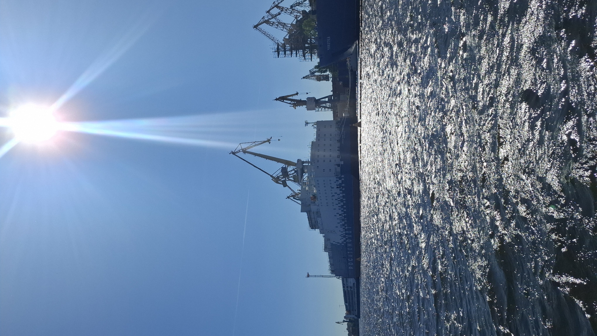 In a sun-drenched Saint Petersburg, Russia, a vast, bright sun shines down from a clear blue sky, its rays creating a visible beam of light. Below, the sunlit, choppy waters of the Neva River reflect the intense sunlight. Anchored in the port are two large vessels. The vessel on the left, a long, white and blue ship, bears the Cyrillic text "РОСАТОМФЛОТ" (Rosatomflot). This ship is accompanied by a smaller, similarly styled vessel docked alongside its stern. Multiple large cranes, some yellow and some darker in color, tower over the ships, indicating ongoing work or construction in the shipyard. To the right, a second, darker blue vessel is docked, with the Cyrillic text "ЧУКОТКА" (Chukotka) clearly visible on its side. The overall scene captures a bustling industrial port under a brilliant, clear sky.
