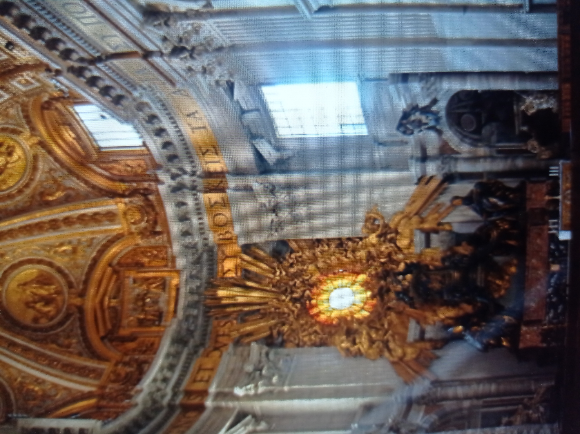 The image depicts the interior of a grand, ornate building with elaborate golden decorations and intricate architectural details. A large, glowing circular object, possibly a stained-glass window or a symbolic representation, is centrally located, surrounded by golden sculptural elements that resemble rays of light and possibly angelic figures. In the foreground, dark, sculpted figures are visible on a pedestal. The walls are lined with tall columns and arches, and there are visible windows with white light. The overall impression is one of religious or historical significance, characterized by opulence and artistry. The provided context suggests this location is in Mukachevo, Ukraine.