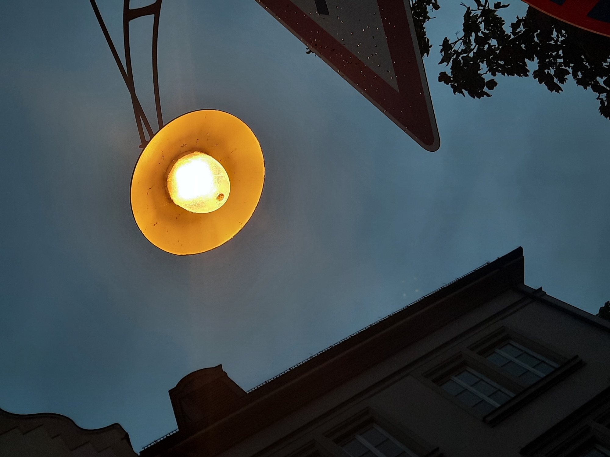 The image captures a low-angle view of a street lamp illuminating the twilight sky over Rosenheim, Germany. The street lamp, with its yellow reflector and visible bulb, is centrally positioned in the upper portion of the frame. To the left, a portion of a building with visible windows is silhouetted against the dusky sky. In the lower right, a triangular traffic sign with red and white markings is partially visible, and a dark silhouette of tree foliage extends from the bottom right corner. The sky is a deep blue-grey, suggesting the transition from day to night. The scene appears to be an outdoor urban setting.